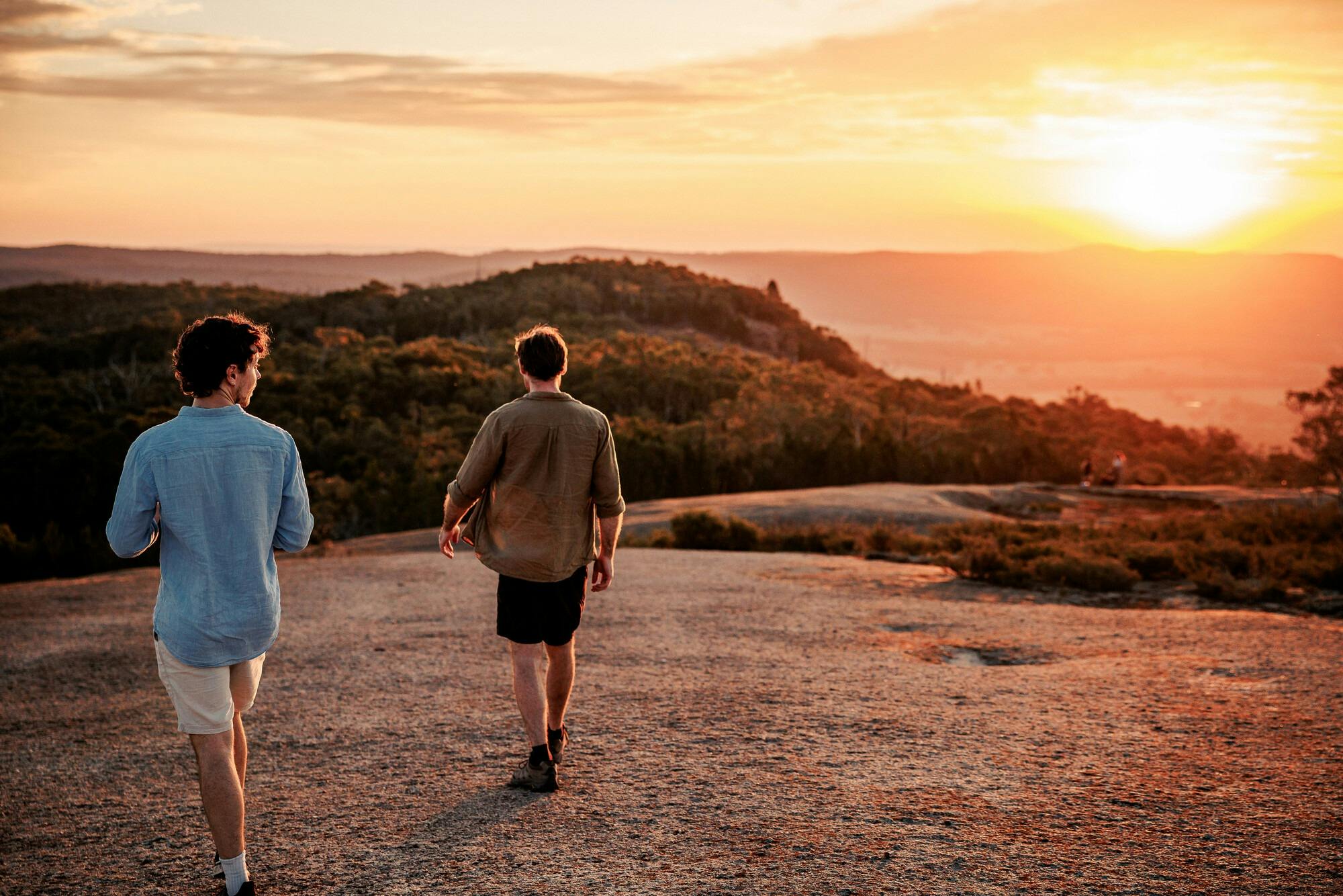 Two men walking on granite plateau at sunset surrounded by hills and bushland
