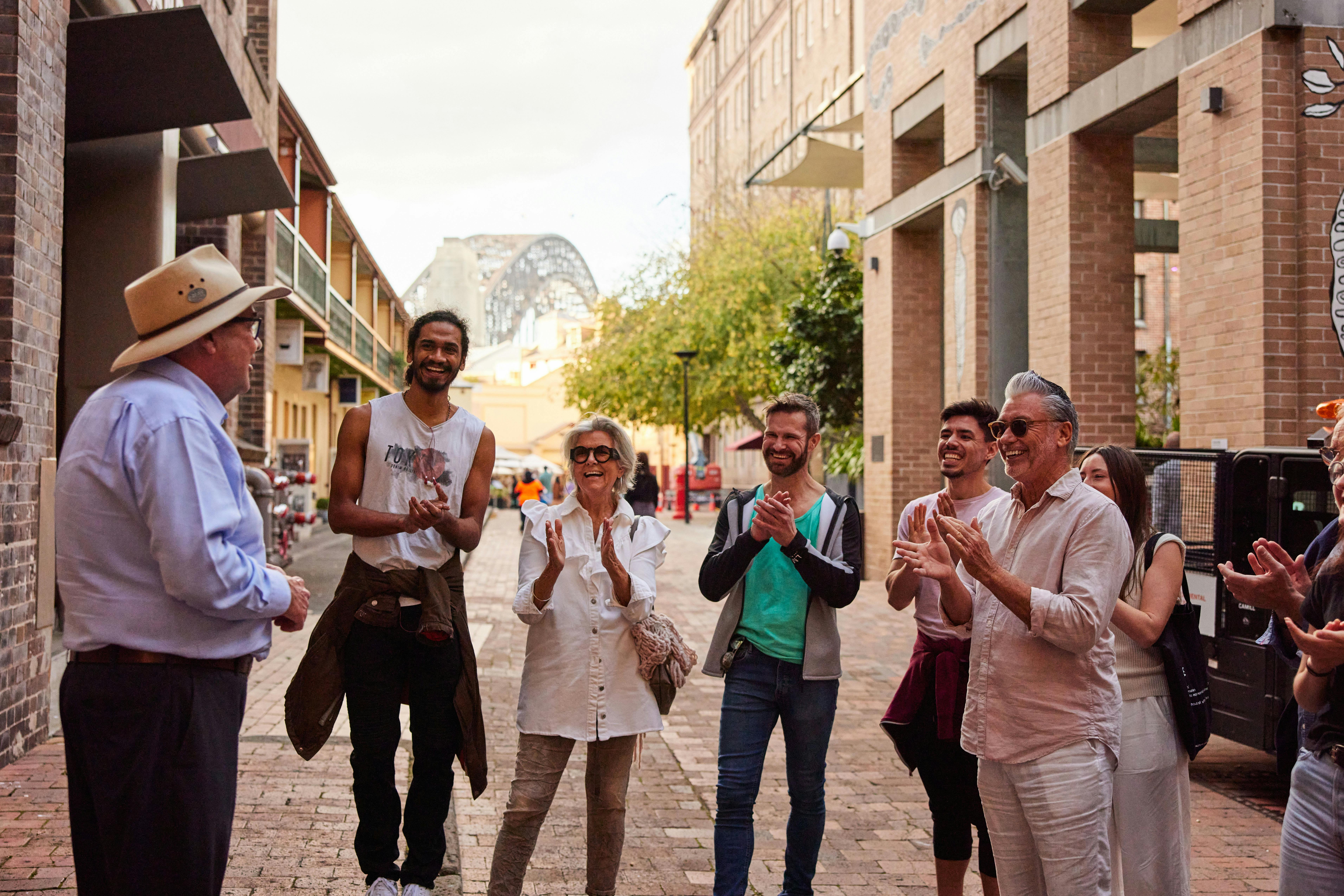 A walking tour guide speaks to a smiling group in a cobblestone laneway in The Rocks, Sydney, with t