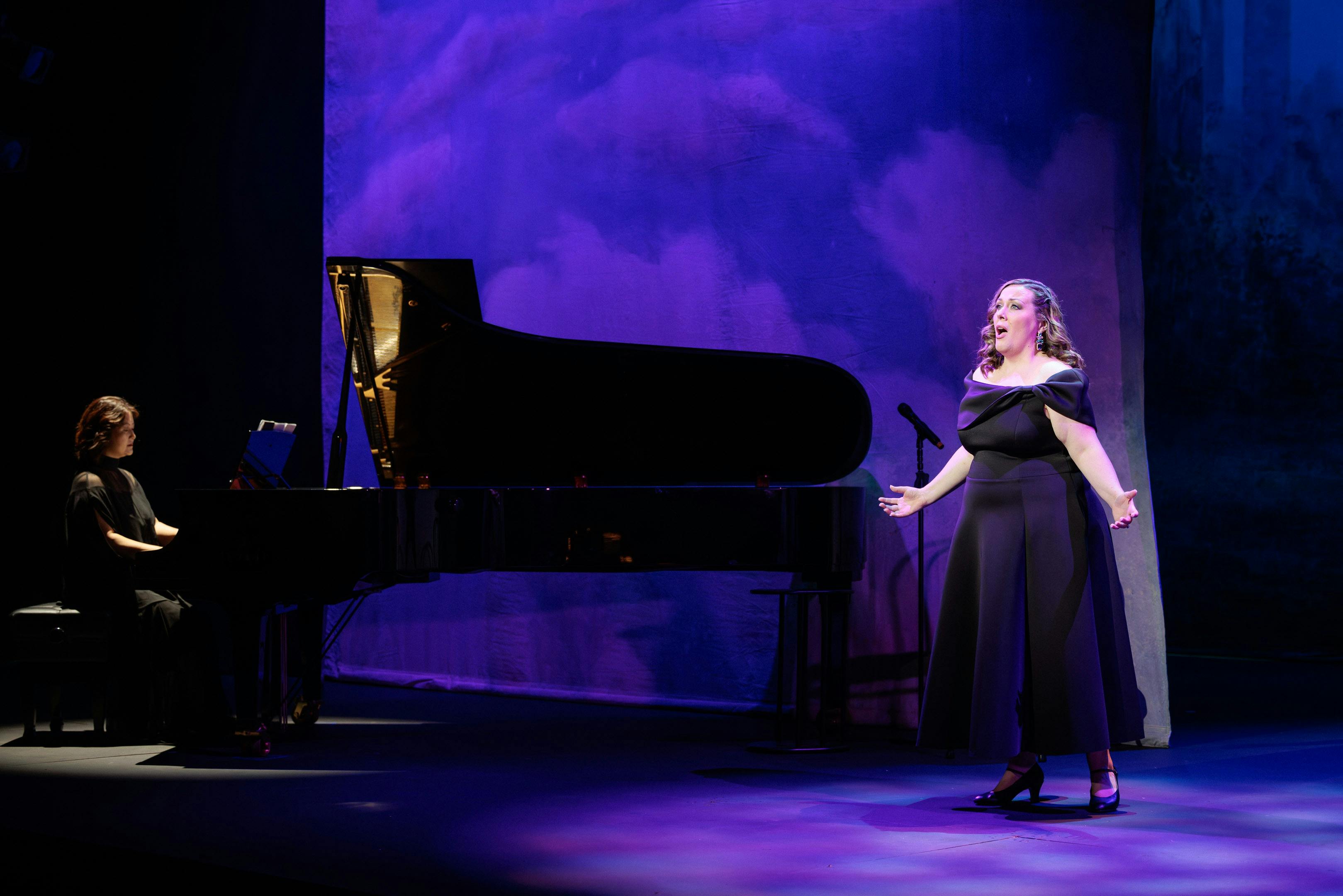 An opera singer in a black dress stands beside a piano singing