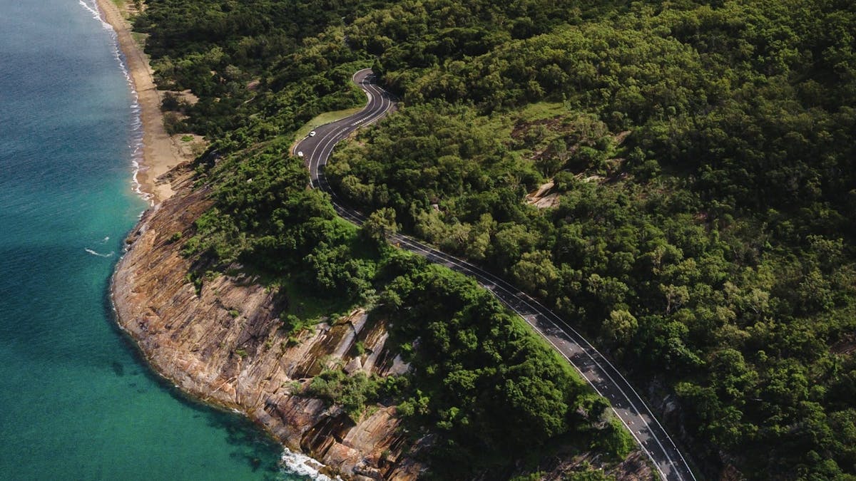 An aerial view shows bush-covered hills and winding roads overlooking the sea below.