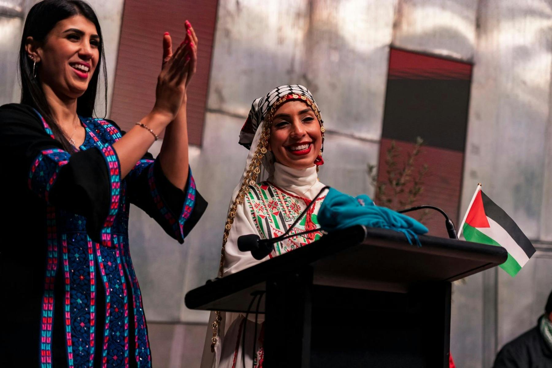 Two women in embroidered Palestinian dresses smiling and clapping on stage beside a lectern