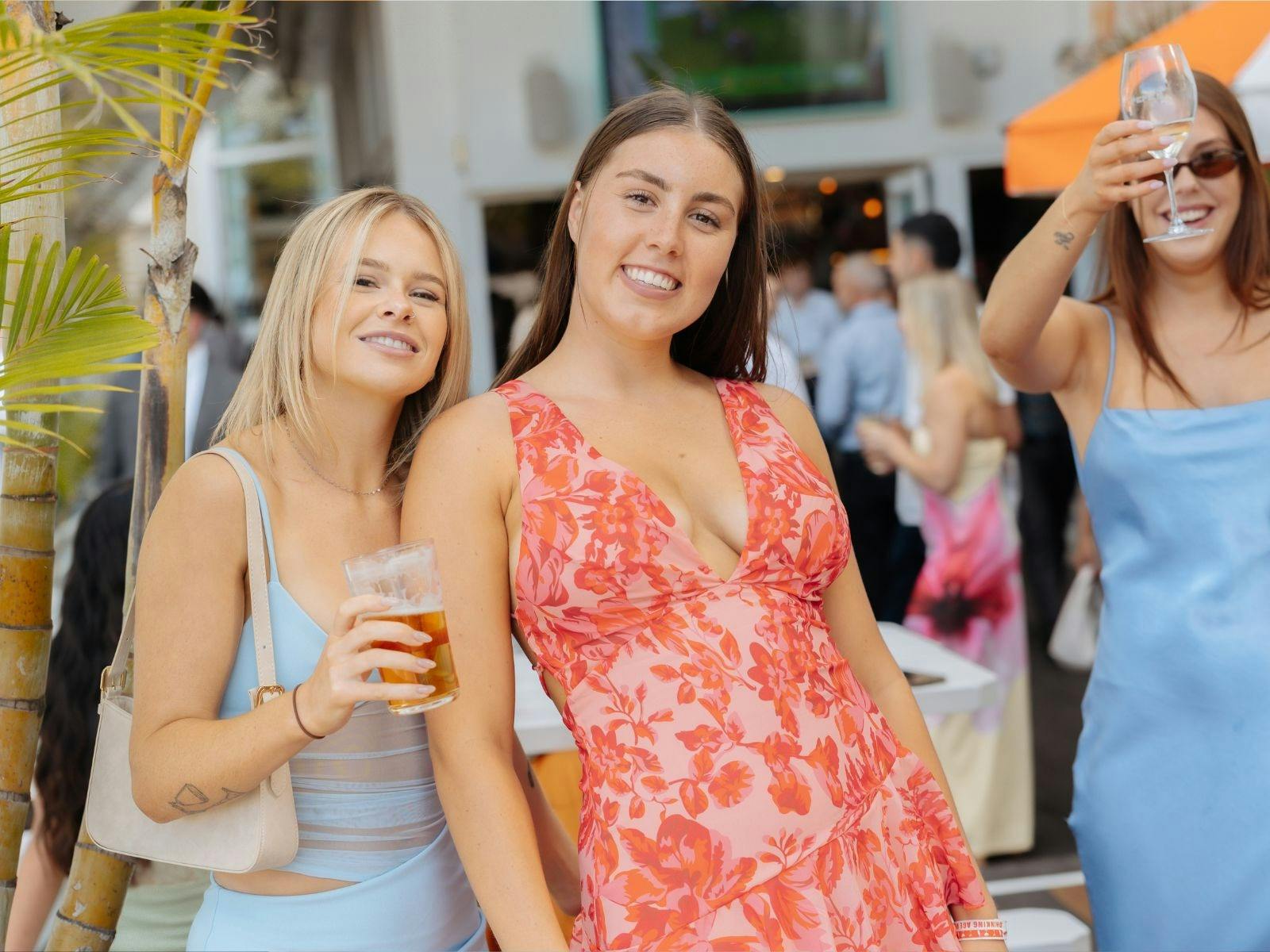 Two women dressed for Melbourne Cup holding drinks and smiling on the outdoor deck at Pepe’s on the
