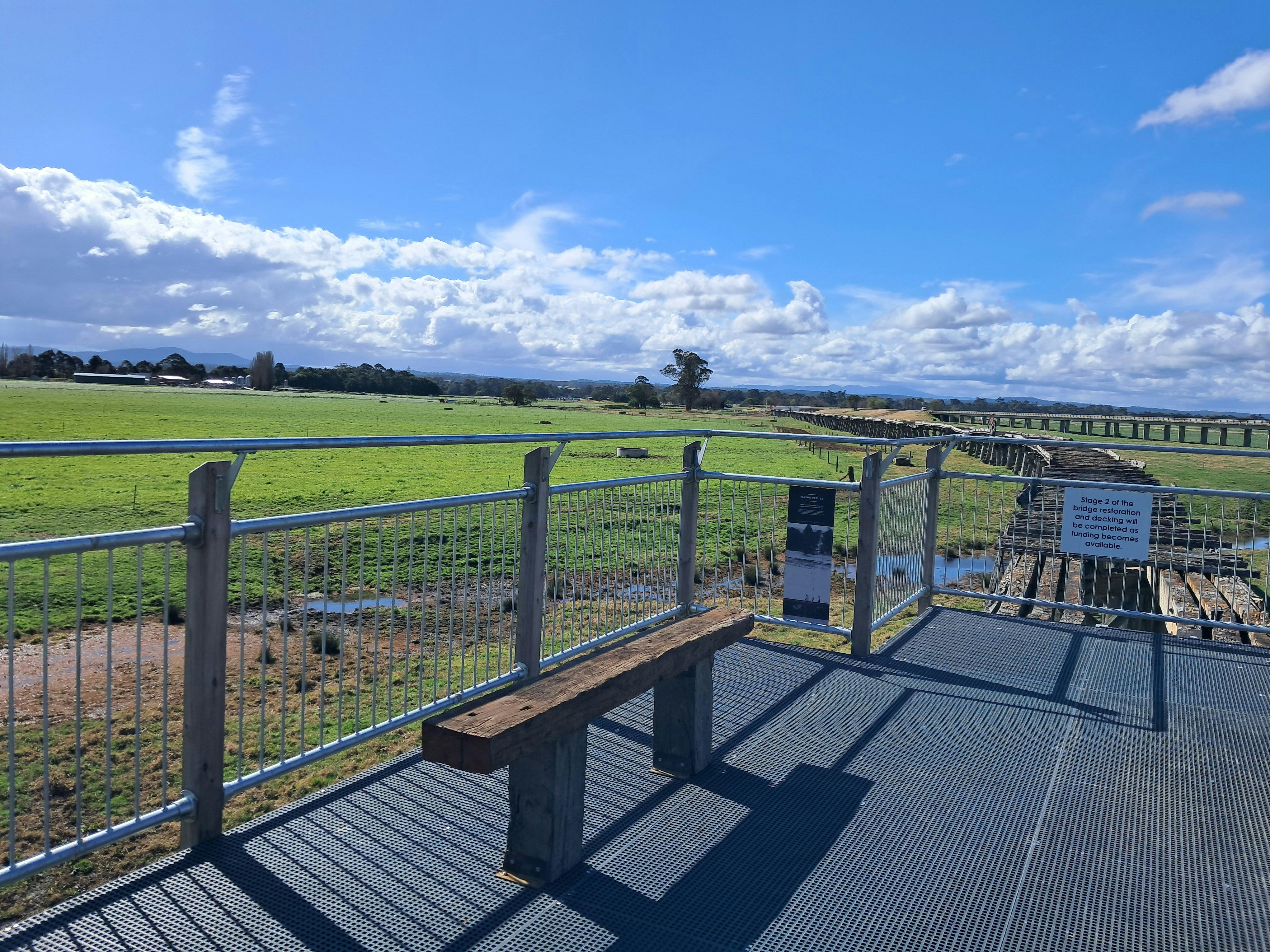 A rustic timber seat on elevated mesh walkway overlooking farmland and historic timber bridge
