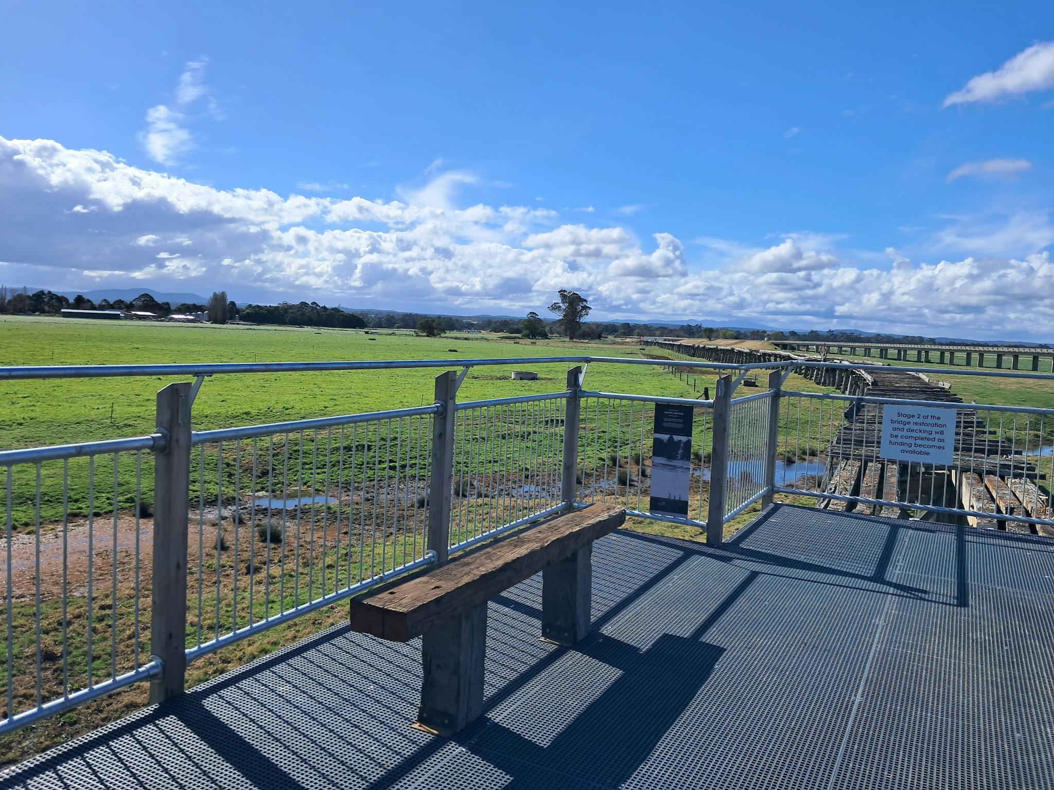 A rustic timber seat on elevated mesh walkway overlooking farmland and historic timber bridge