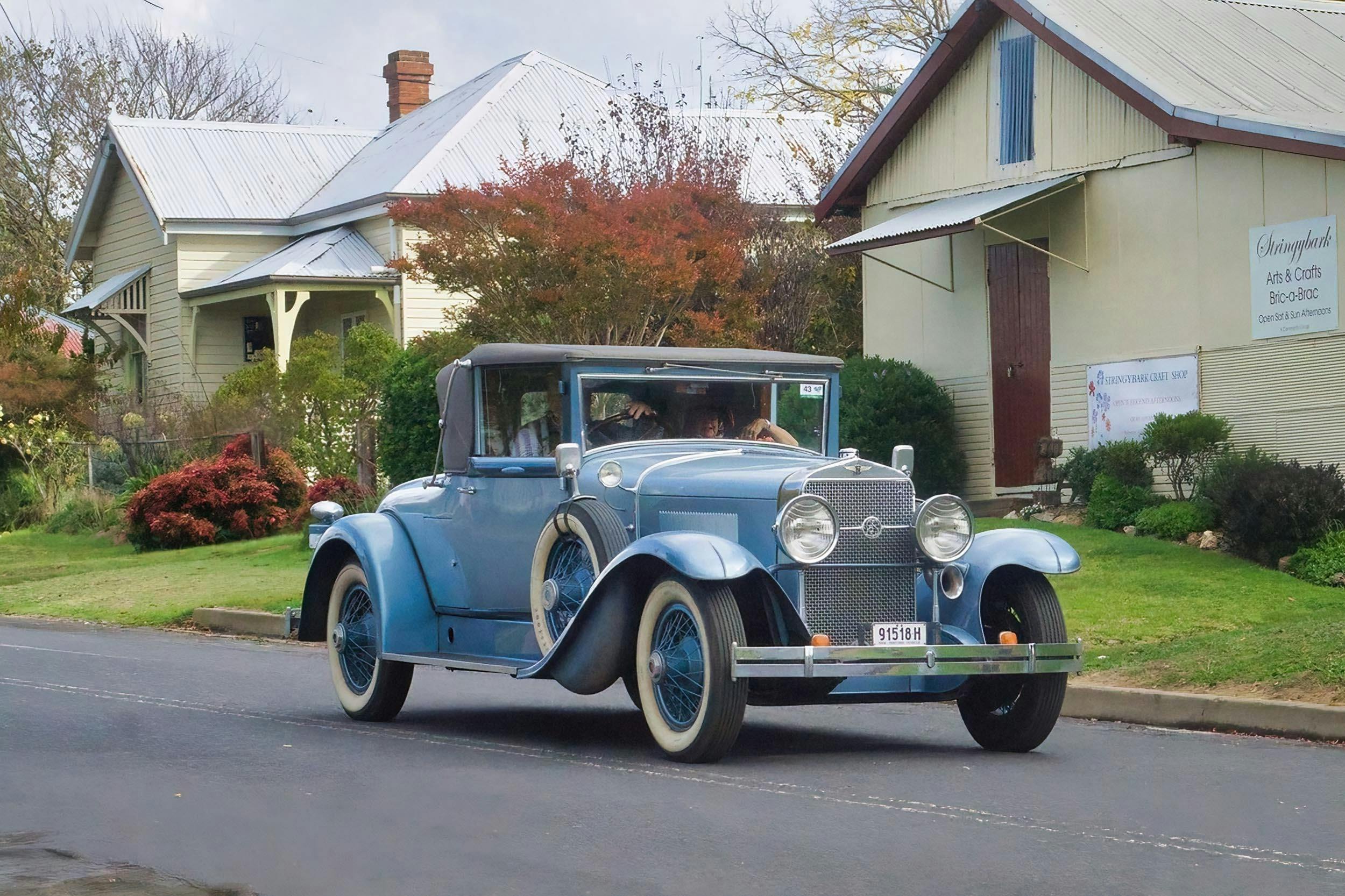 A blue vintage car is being driven past the cream fibro craft shop and a cream weatherboard house