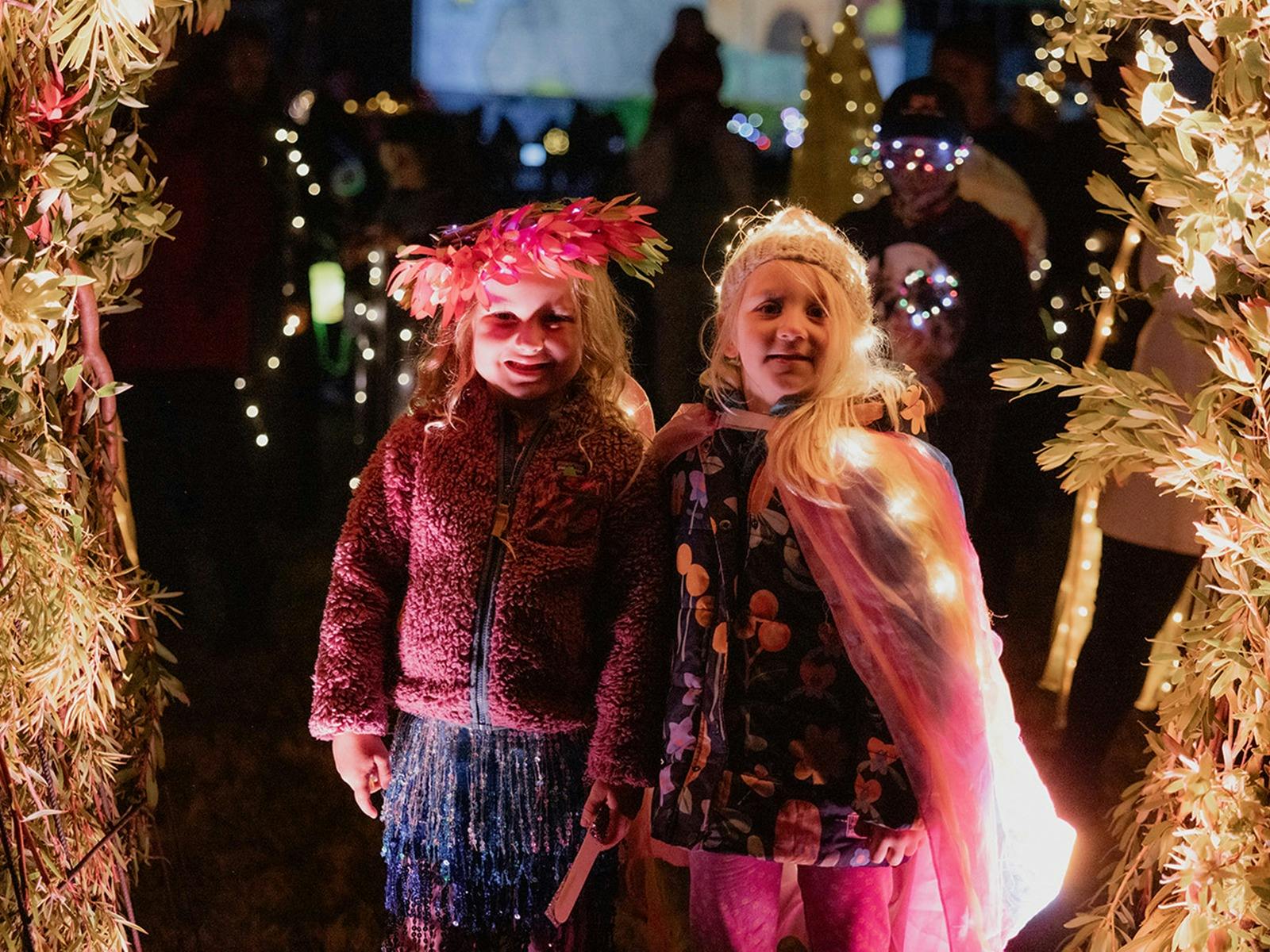 Two young people wearing headdresses and capes with fairy lights, pose under a floral archway.