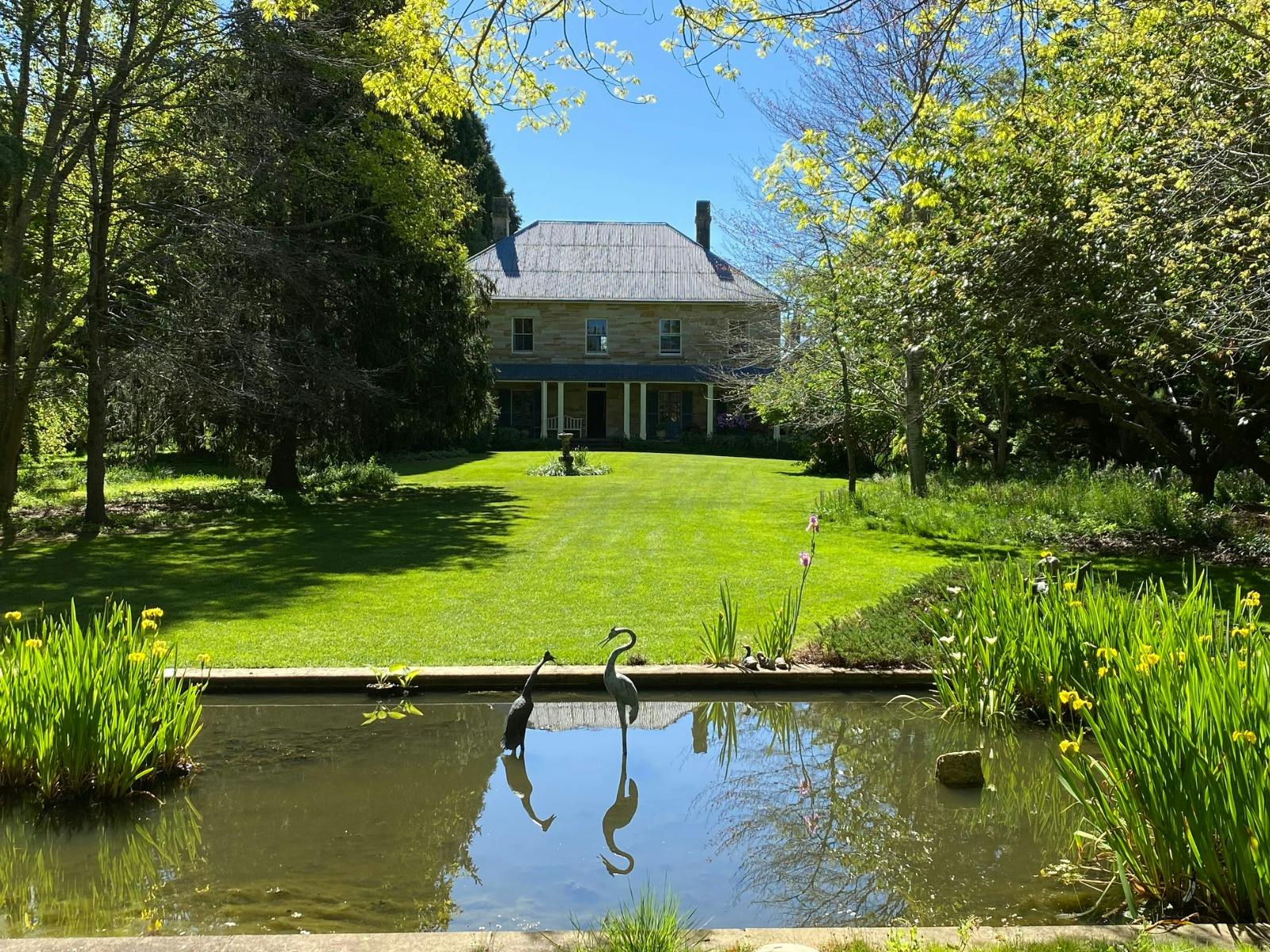 Historic sandstone double storey building overlooking a grassy lawn. A small pond with stalk statues