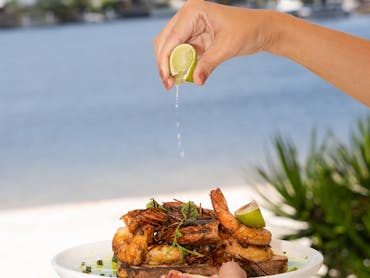 Hand squeezing lime onto grilled prawns on a toasted garlic sourdough bread with a view of the river