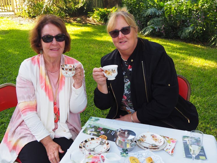 Two women sit side by side holding a cup of tea with cakes and wine on the table in the foreground