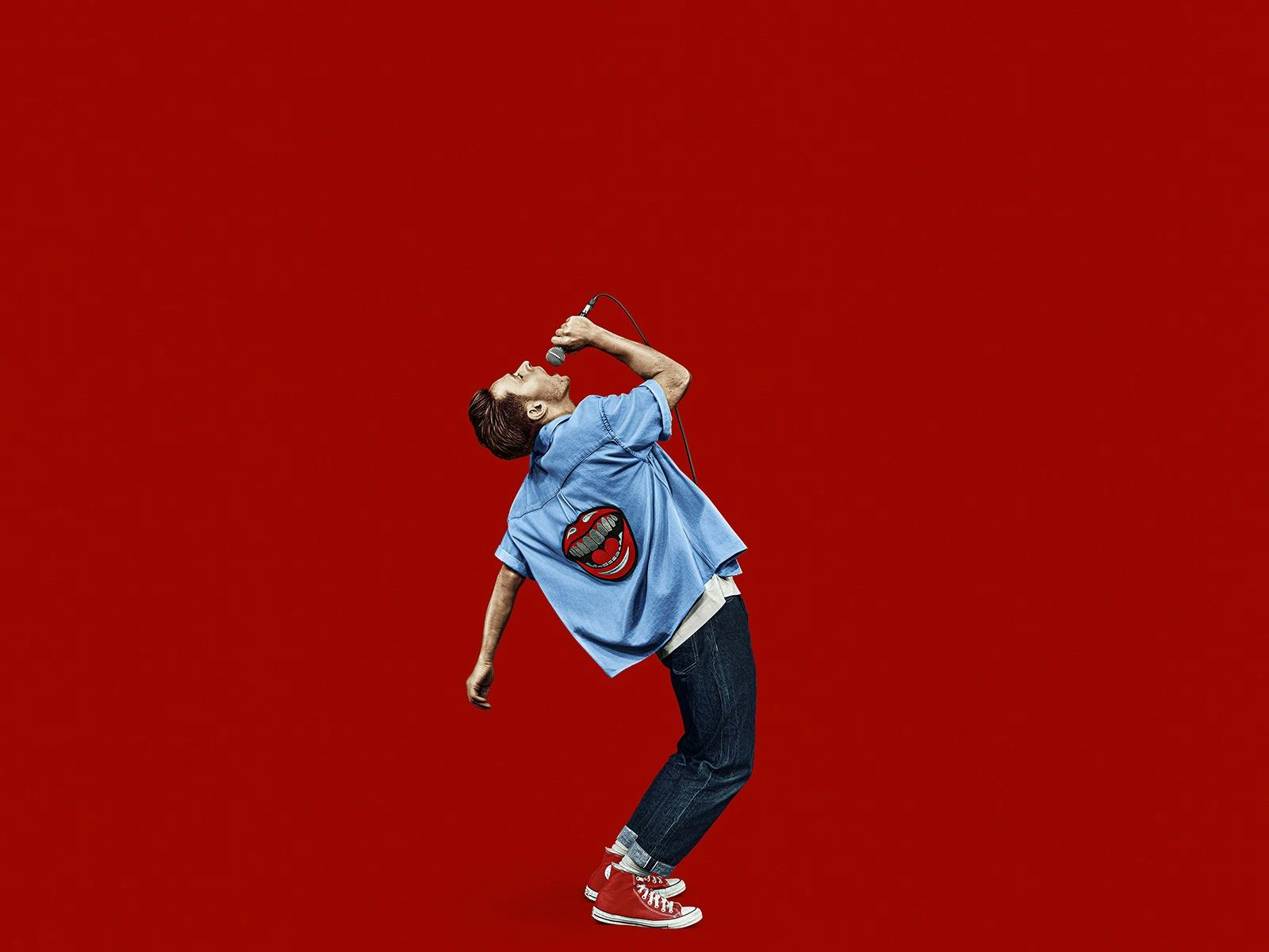 A man stands in front of a red background. He wears a denim jacket and holds a microphone.