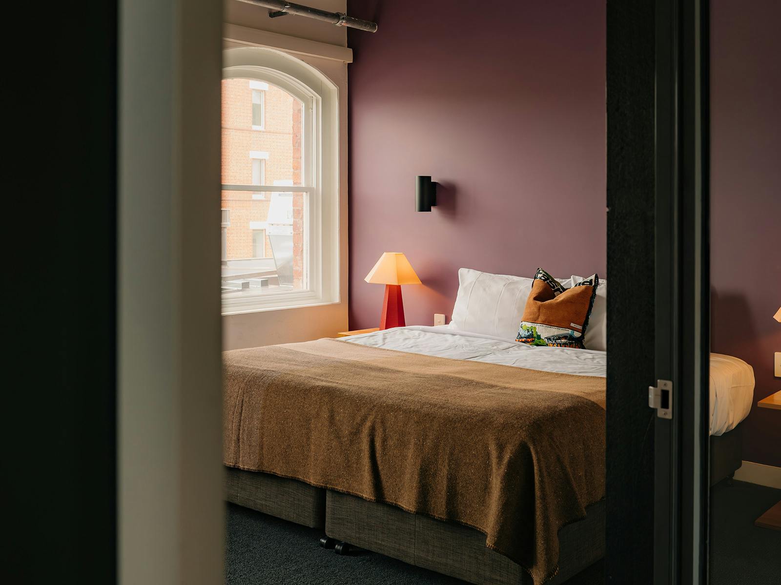 Bedroom with plum-toned wall, made bed, and warm bedside lamp lighting.