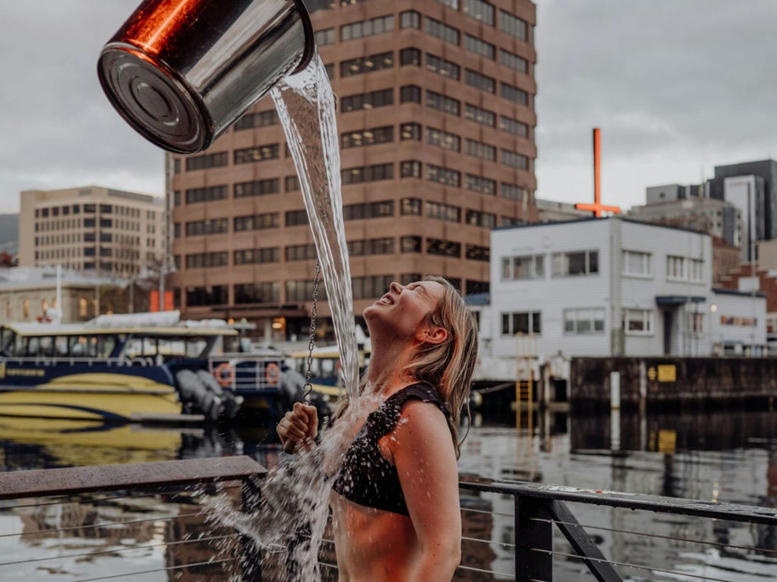 A woman smiles as a sheet of water from a bucket shower pours over her on a pier.