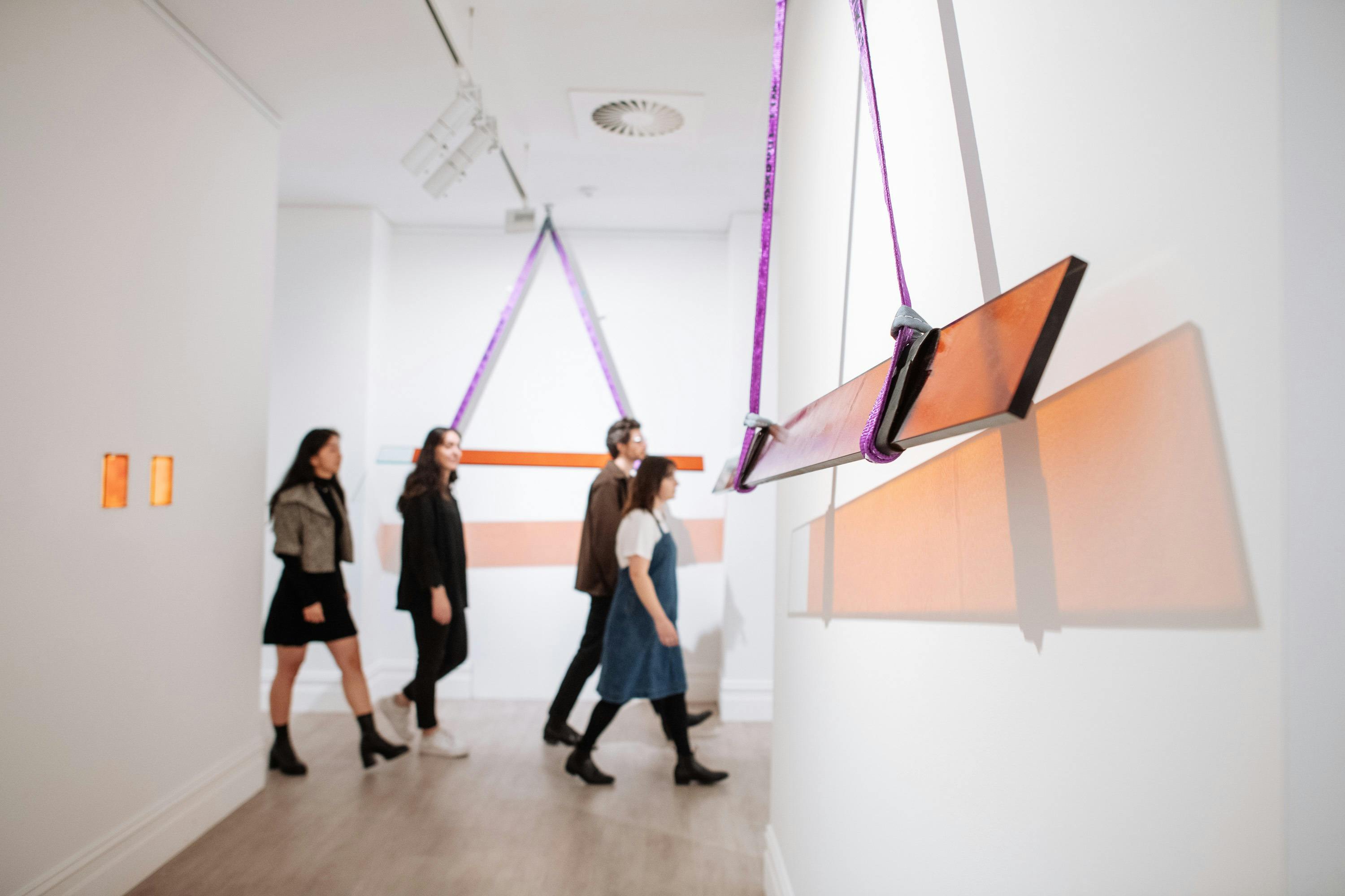 Group of four people walk through a gallery with hanging amber coloured glass panel sculptures