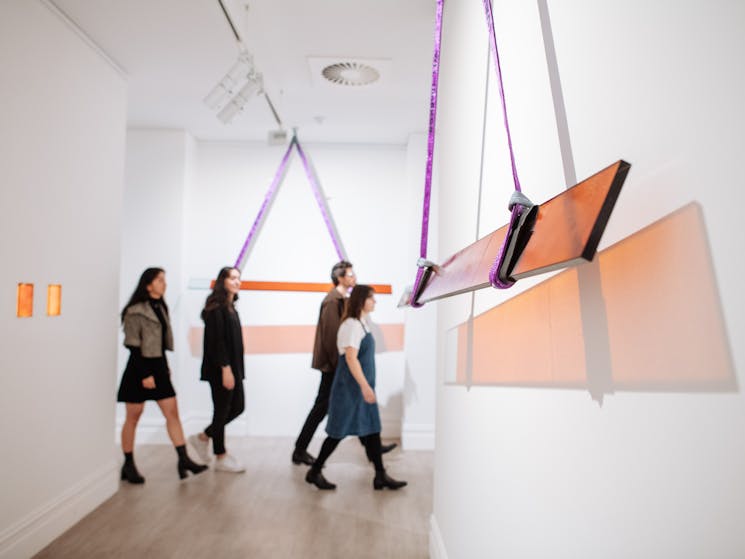 Group of four people walk through a gallery with hanging amber coloured glass panel sculptures