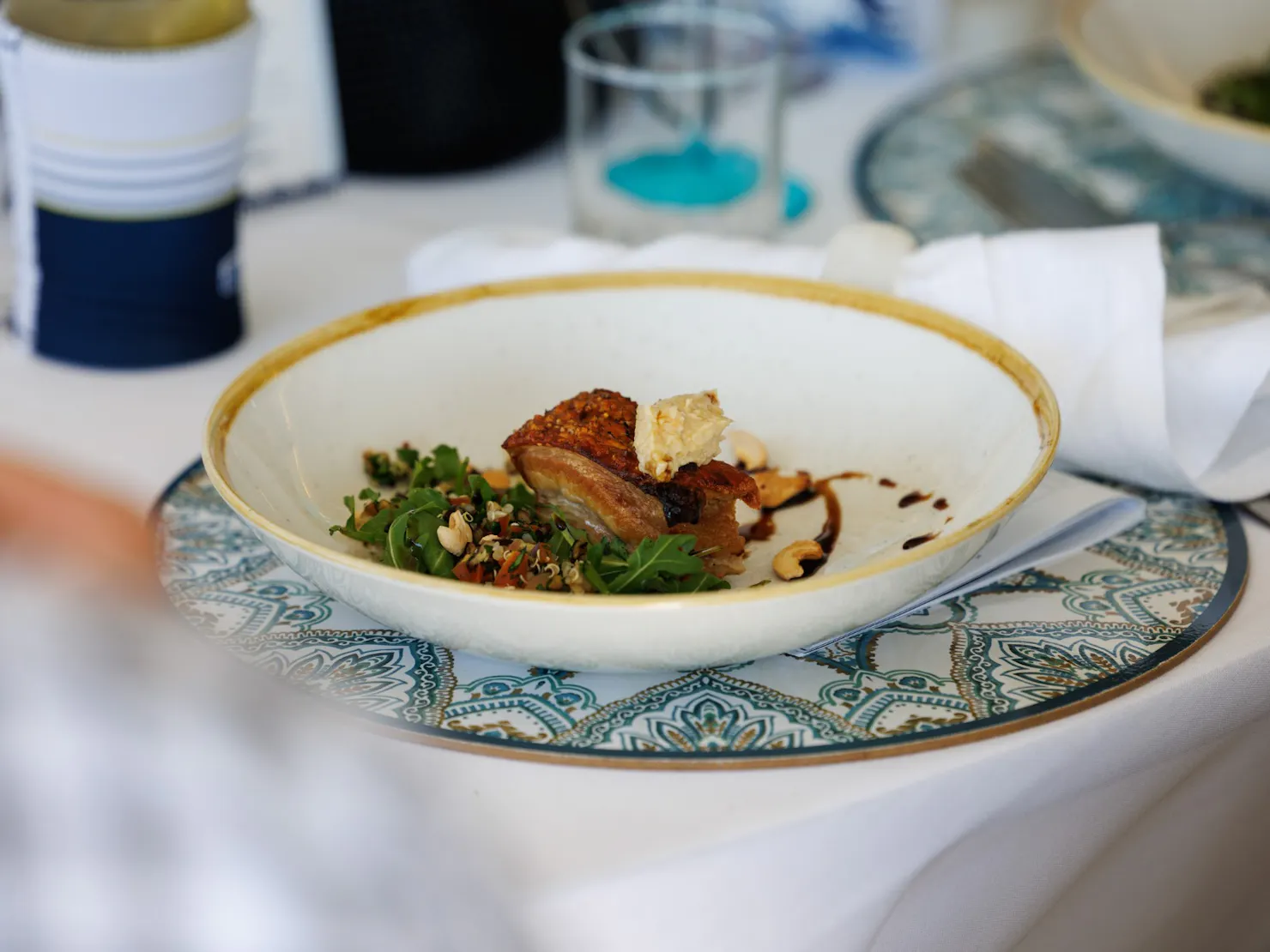 A white bowl on a patterned plate holding a piece of crispy-skinned pork belly with a herb and grai