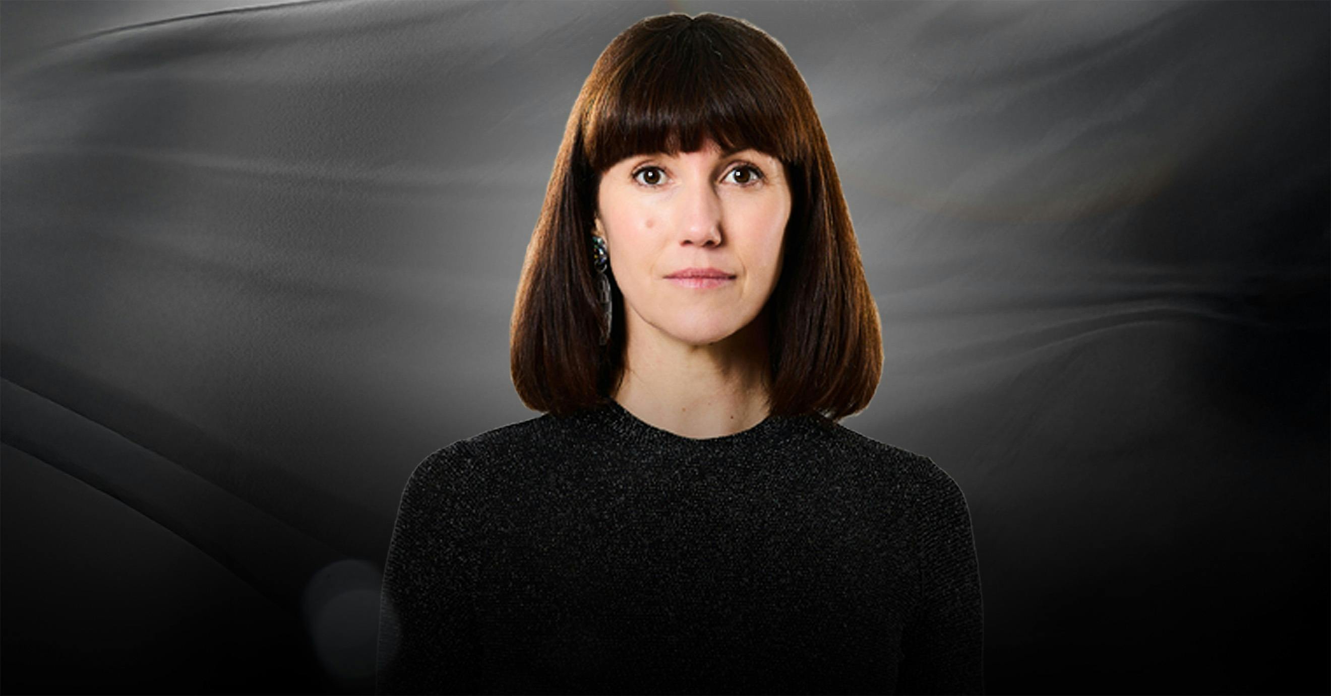 A woman with a long bob and bangs, staring blankly at the camera with a dark background