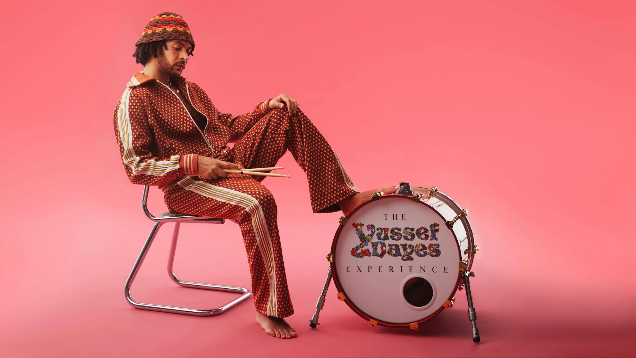 Yussef Dayes - person in beige tracksuit seated on a chair with their foot resting on a kick drum