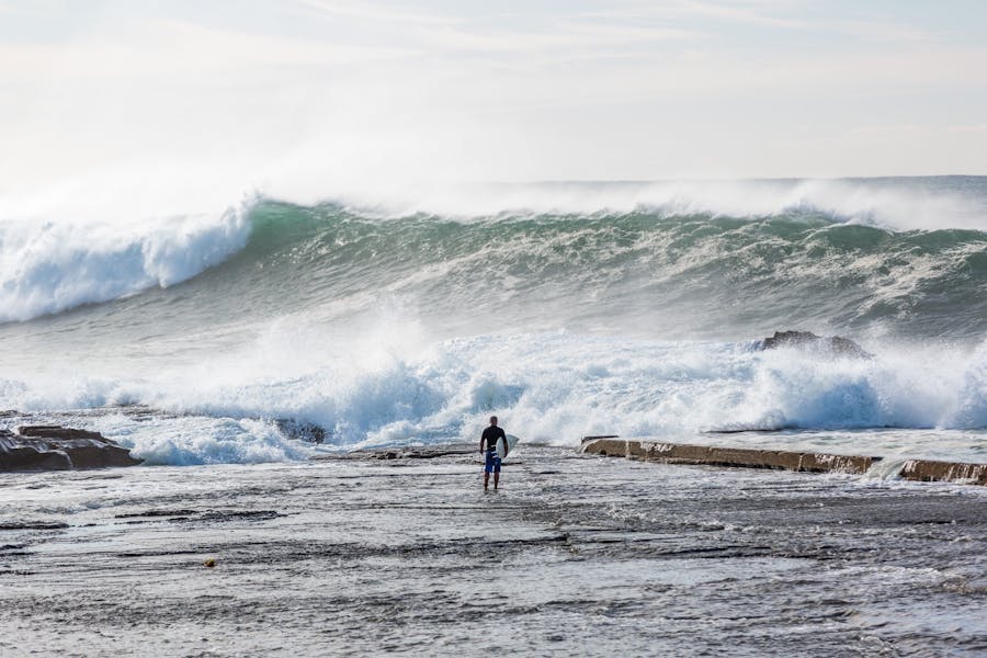 A surfer stands on a rock shelf with big surf rolling in