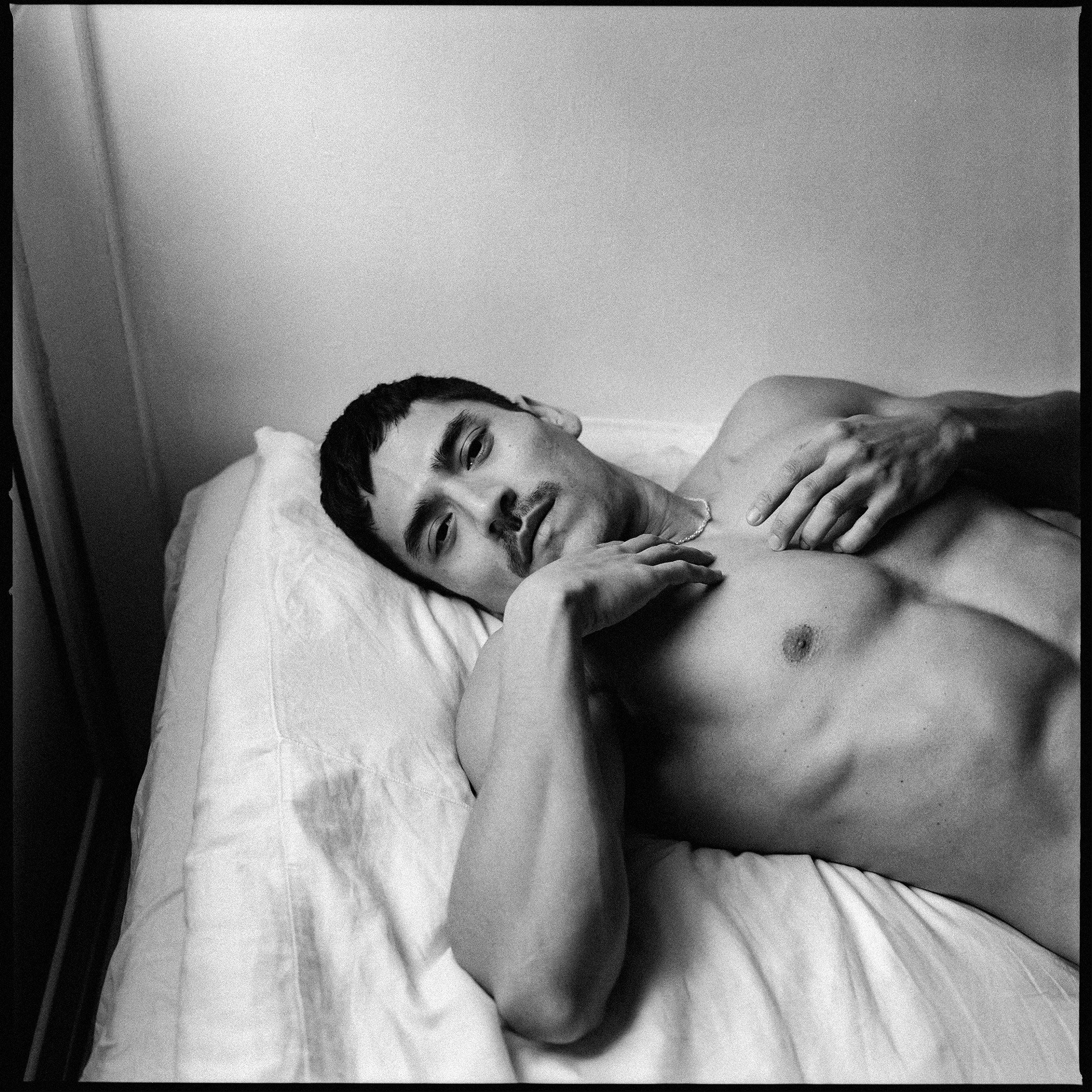 A monochrome pigment photographic portrait of a shirtless man laying on a bed, looking at the camera