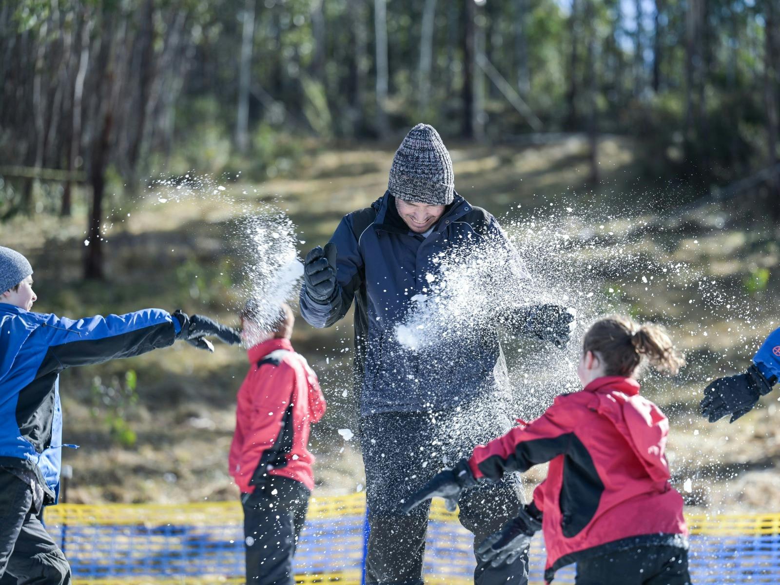 Kids throw snowballs at their dad. Snow is spraying and there is a forest in the background.