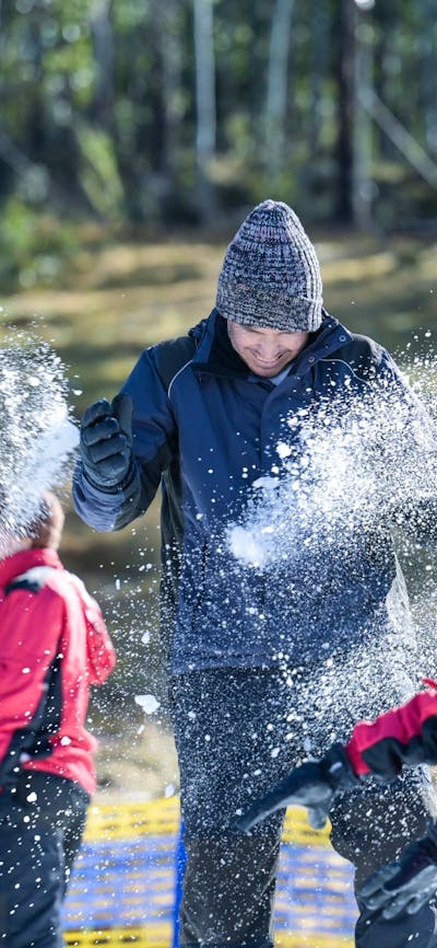 Kids throw snowballs at their dad. Snow is spraying and there is a forest in the background.