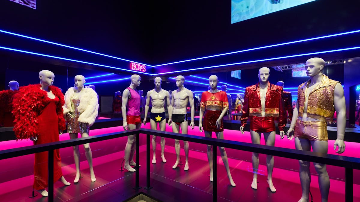 Mannequins dressed in vibrant outfits made of leather, feathers and sequins in a night club setting