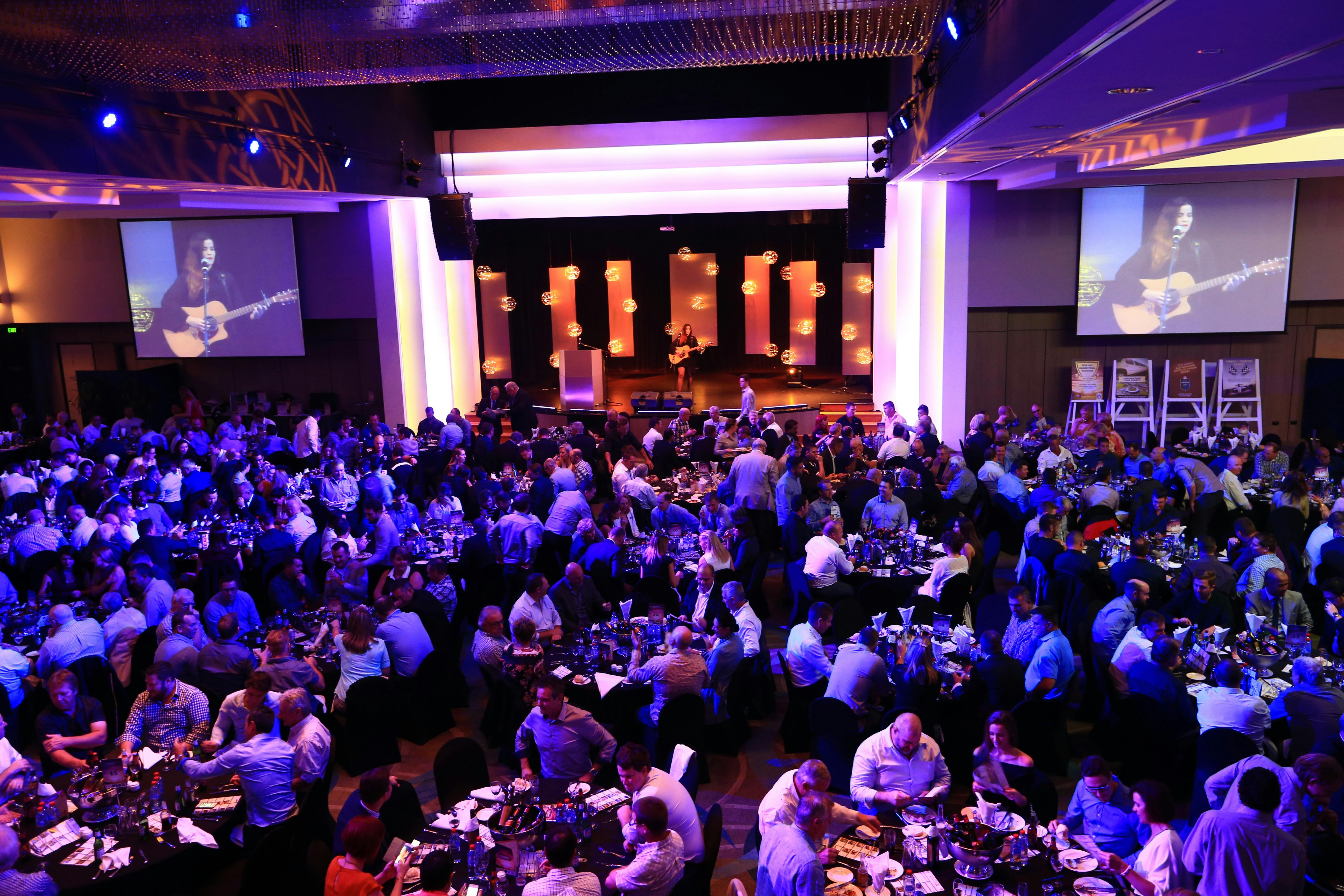 A crowd of people attending an event, seated at tables with a musician on stage