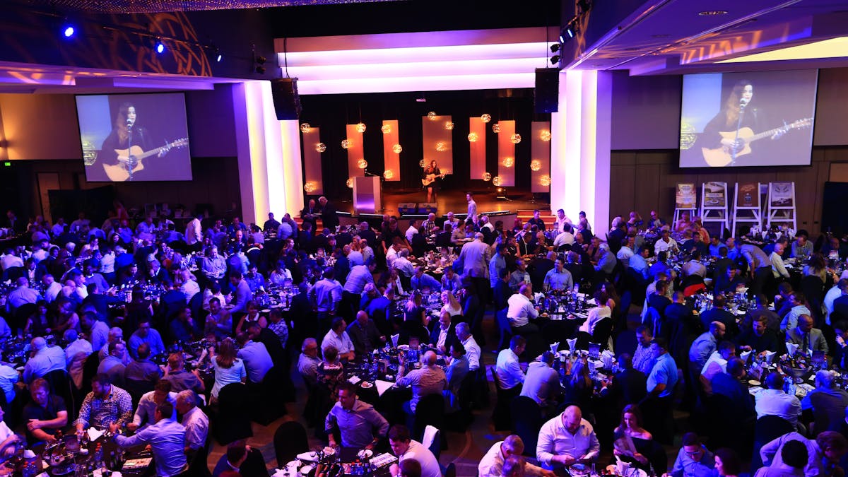 A crowd of people attending an event, seated at tables with a musician on stage A crowd of people attending an event, seated at tables with a musician on stage