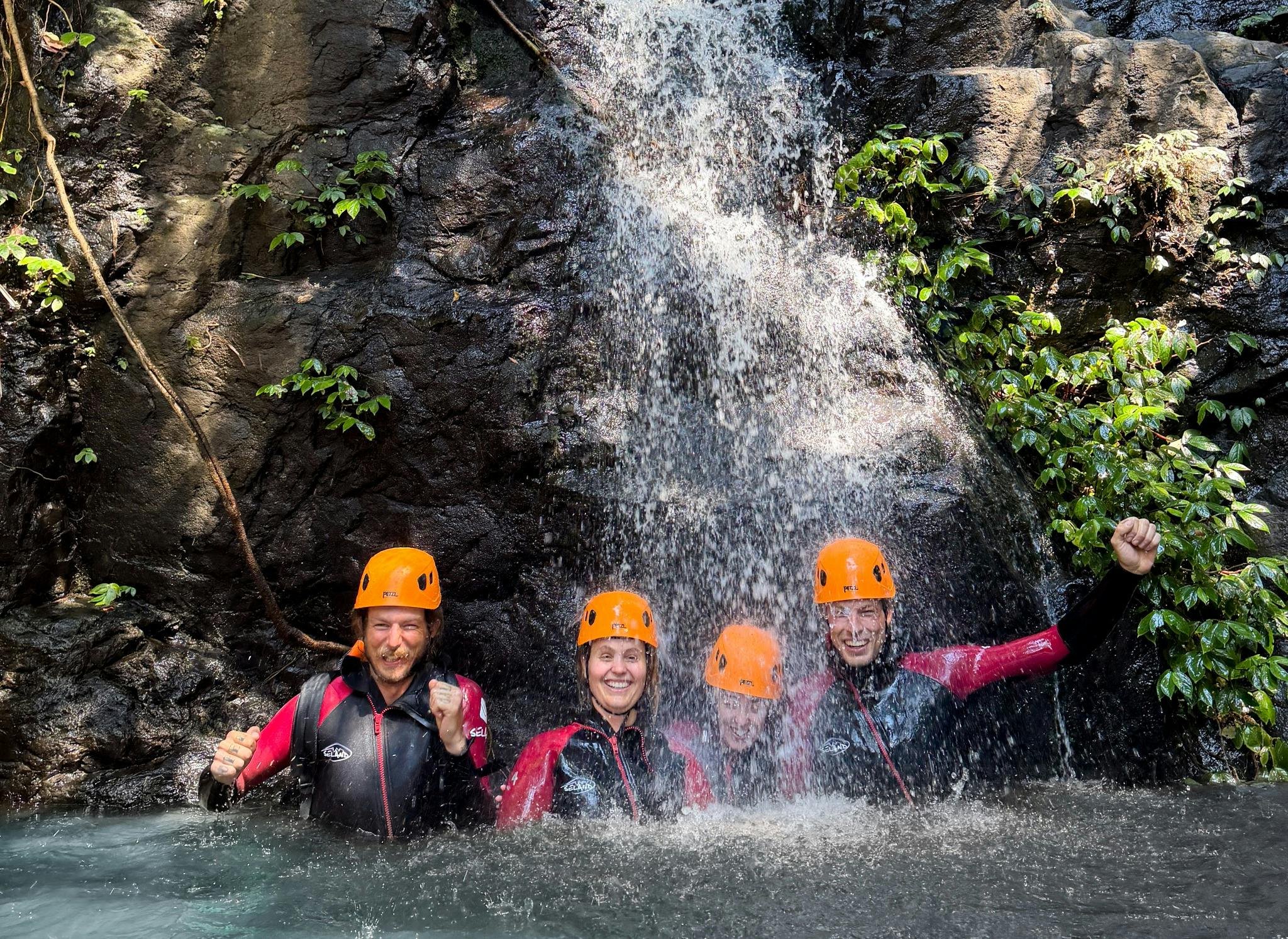 Four canyoners in orange helmets smiling beneath a waterfall in a natural rock pool.