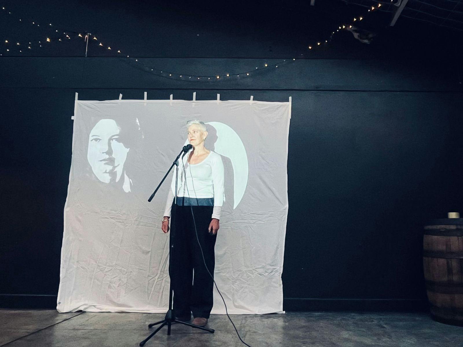 A woman stands in a spotlight next to the projected image of a woman's face