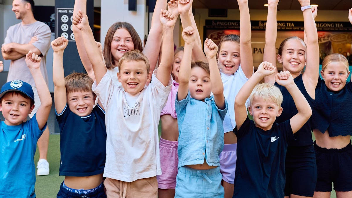 A large group of excited children cheer with fists raised outside, adult smiling in the background. A large group of excited children cheer with fists raised outside, adult smiling in the background.