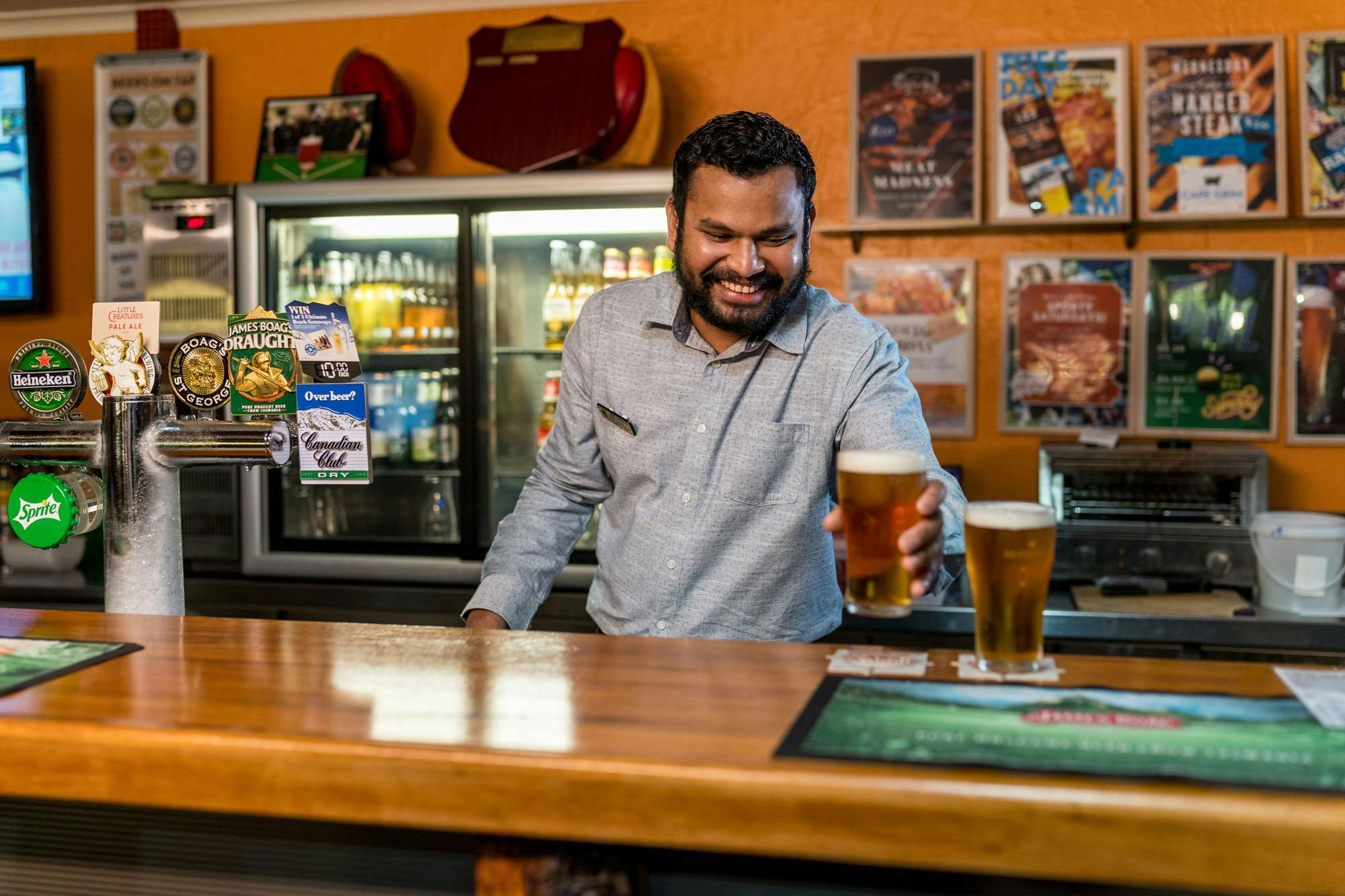Kasun serving beers at the bar with a friendly smile. The 7 Days 7 Ways offer posters on the wall.