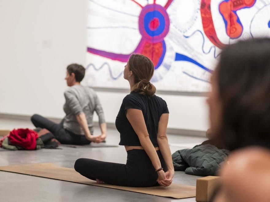 Three participants sit on their yoga mat in the Art Museum stretching with paintings behind