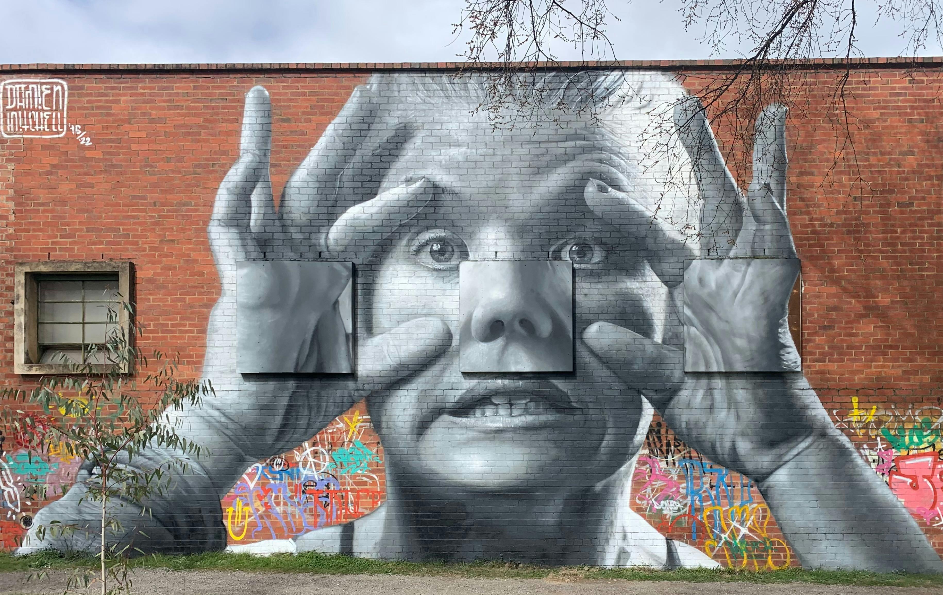 Mural of a face peering out curiously, with hands holding eyes wide. Painted by Damien Mitchell