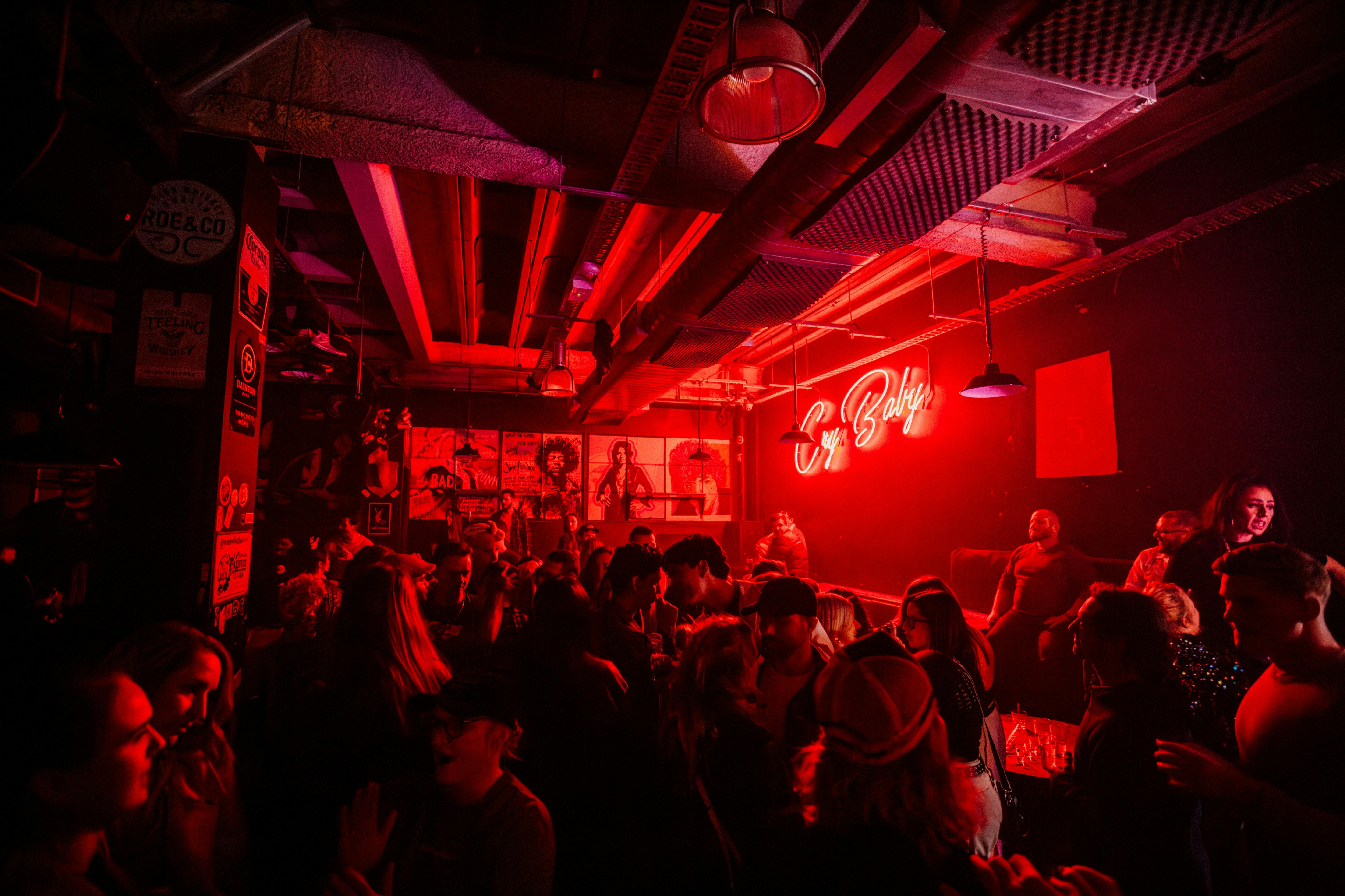 Dark venue, bright pink neon sign 'Cry Baby' on the wall, crowd of patrons socialising