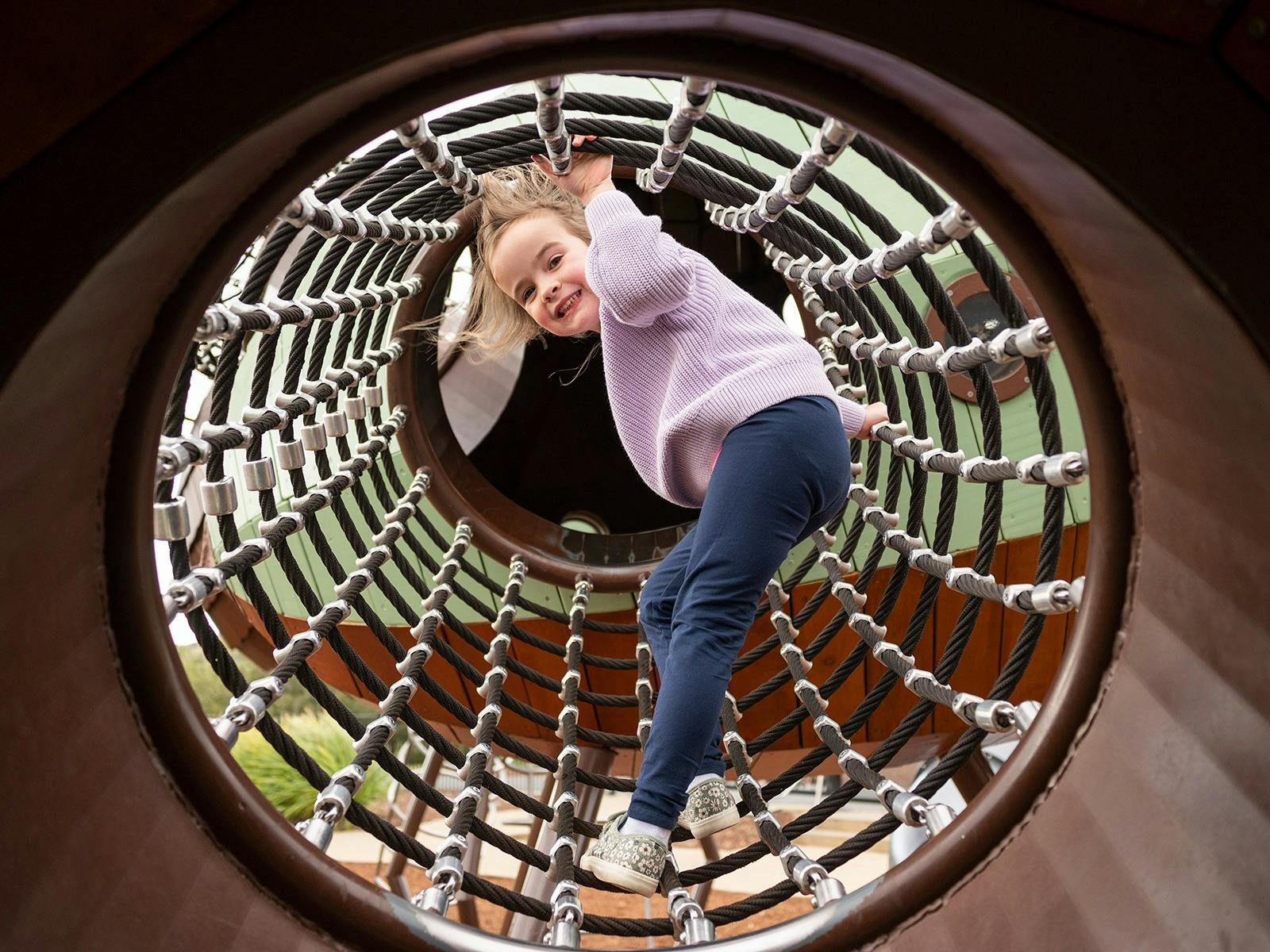 Young girl smiling at camera as she climbs through a net tunnel