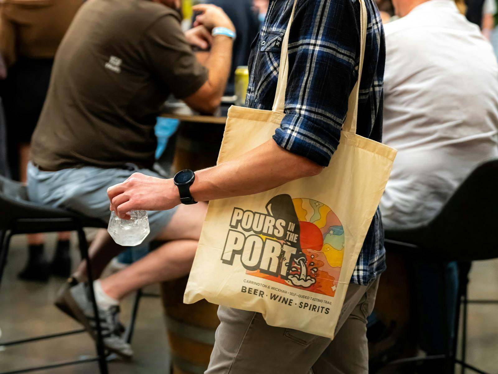 Framed at the shoulder and hip of a festival goer, we see the Pours In The Port tote & sample glass