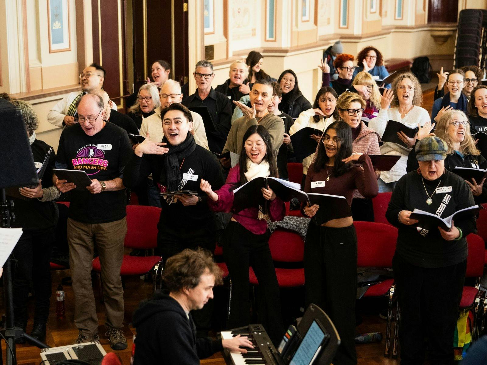 Diverse adult choir singing enthusiastically, holding music books, with a pianist.