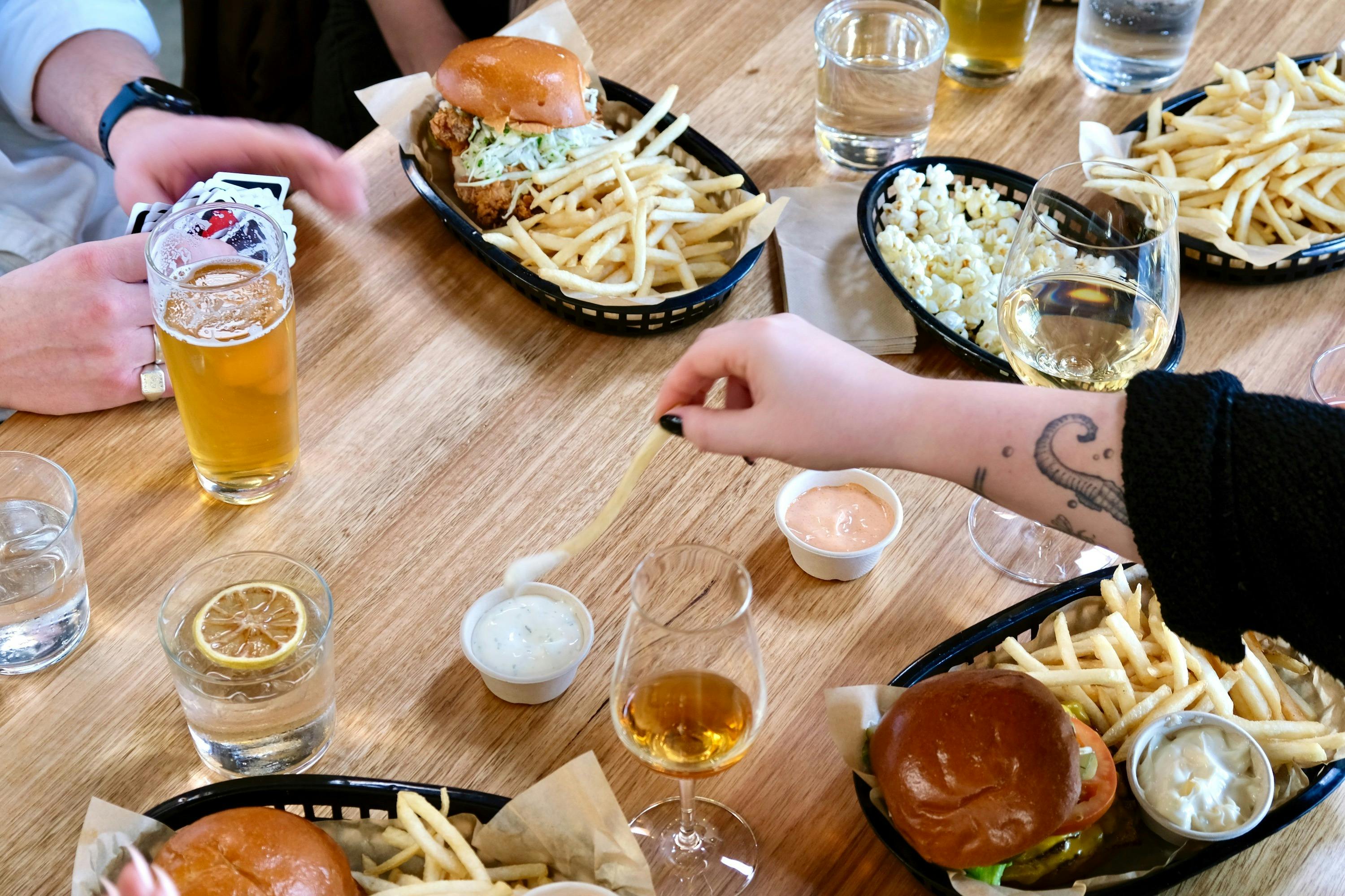 Hand reaching to dip a french fry over tablescape of  baskets of burgers, fries, beer, whisky, wine