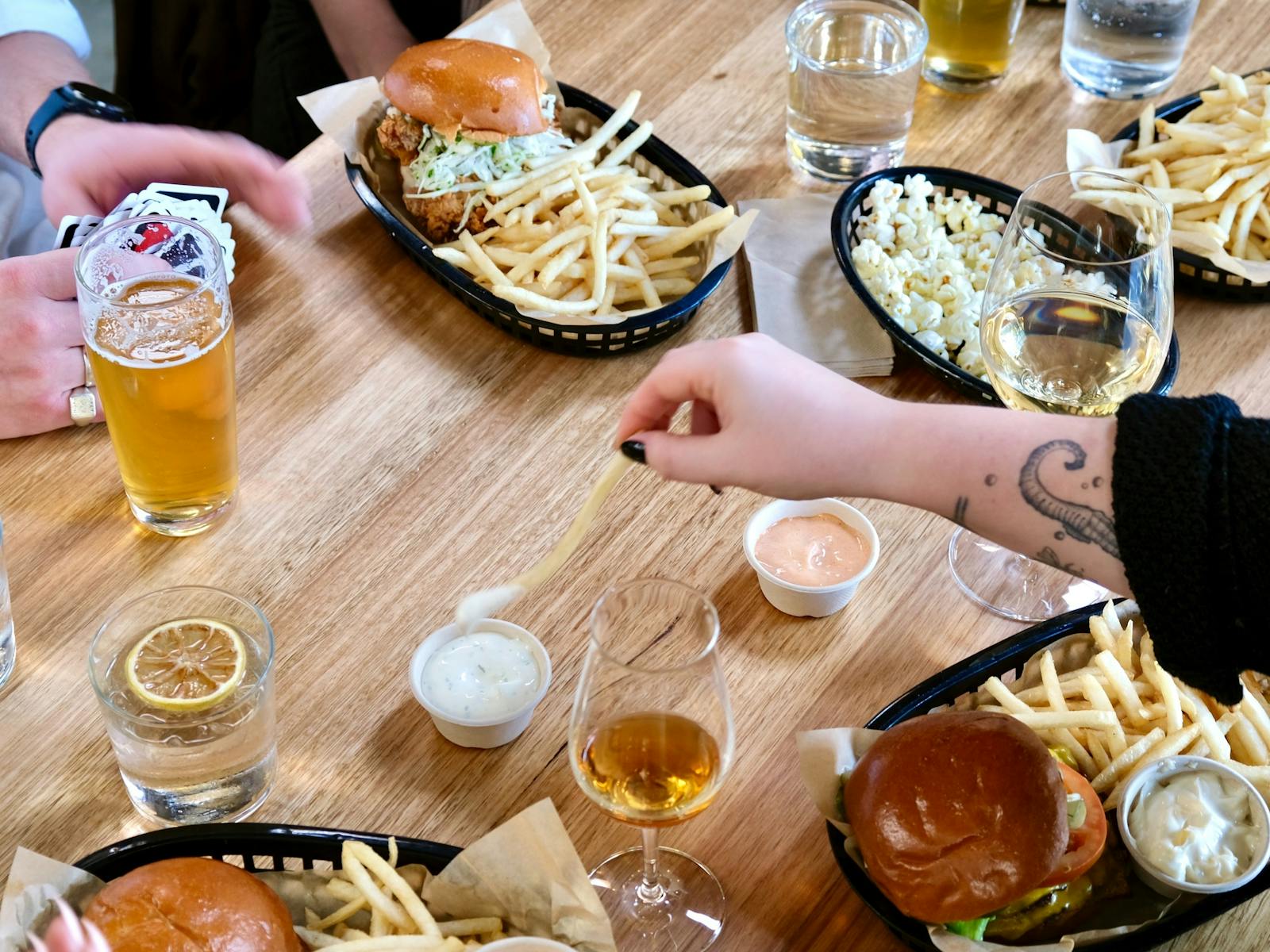 Hand reaching to dip a french fry over tablescape of  baskets of burgers, fries, beer, whisky, wine
