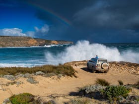 A 4WD parked at the edge of a rocky beach where a wave breaks down on the beach