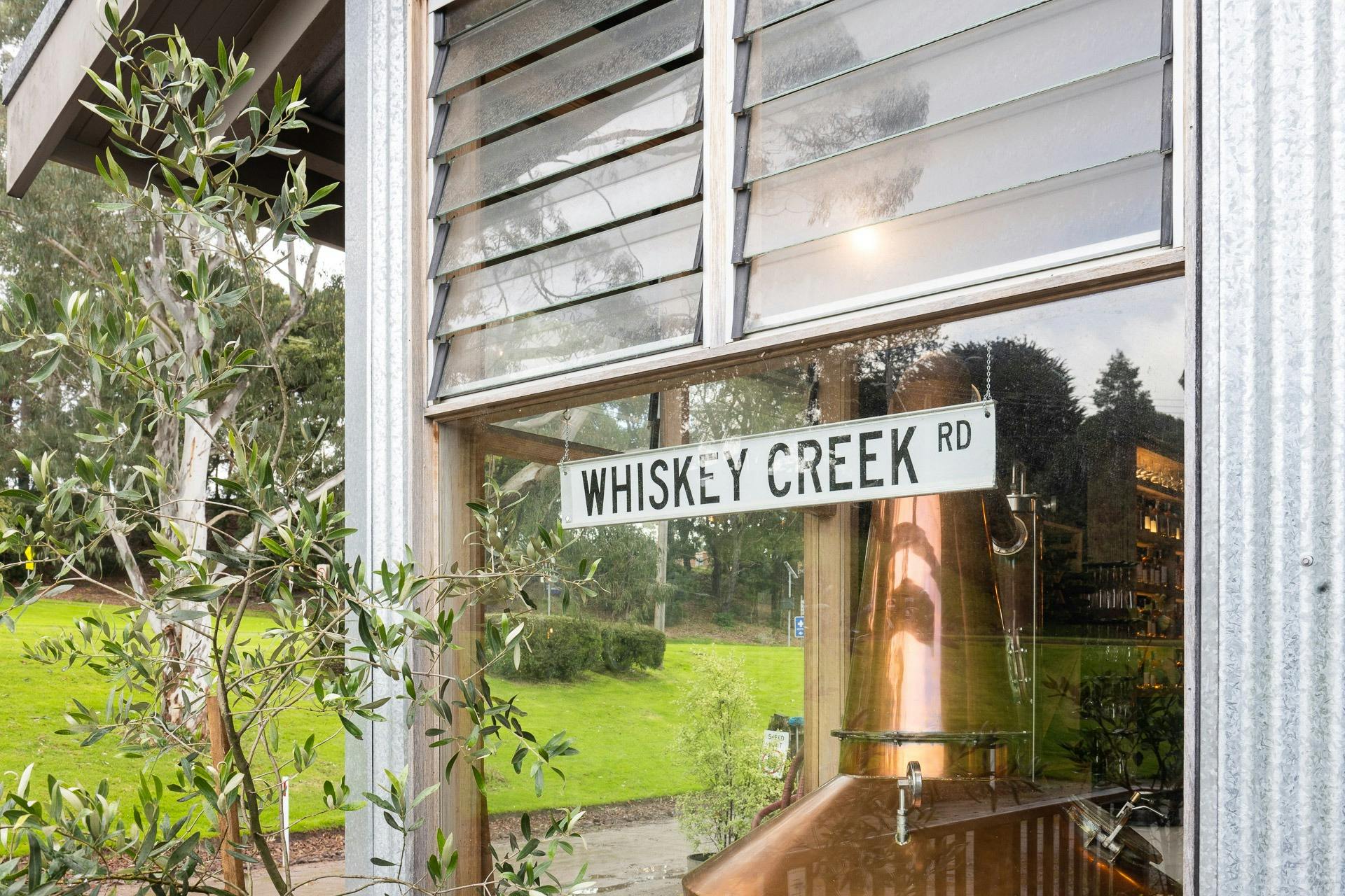 A copper still seen through a window, an old street sign is on the window reads "Whisky Creek Rd"