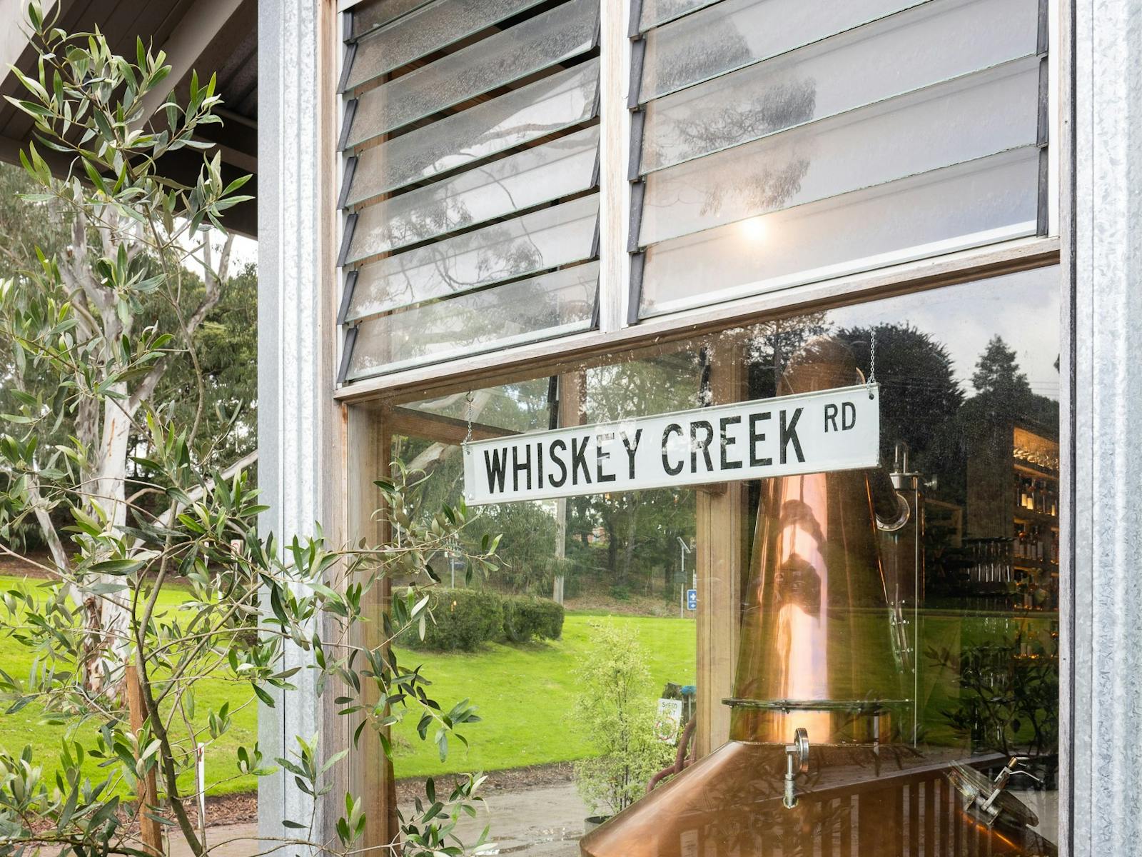 A copper still seen through a window, an old street sign is on the window reads "Whisky Creek Rd"