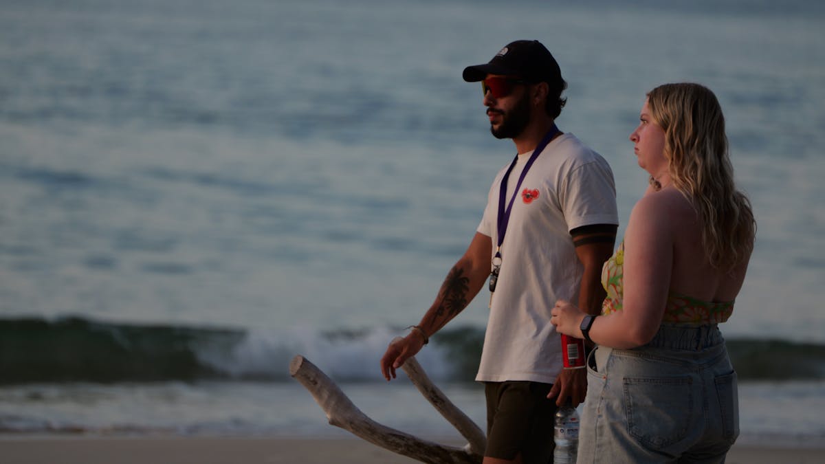 Two people walking side by side on the beach at dusk, with waves in the background and warm evening