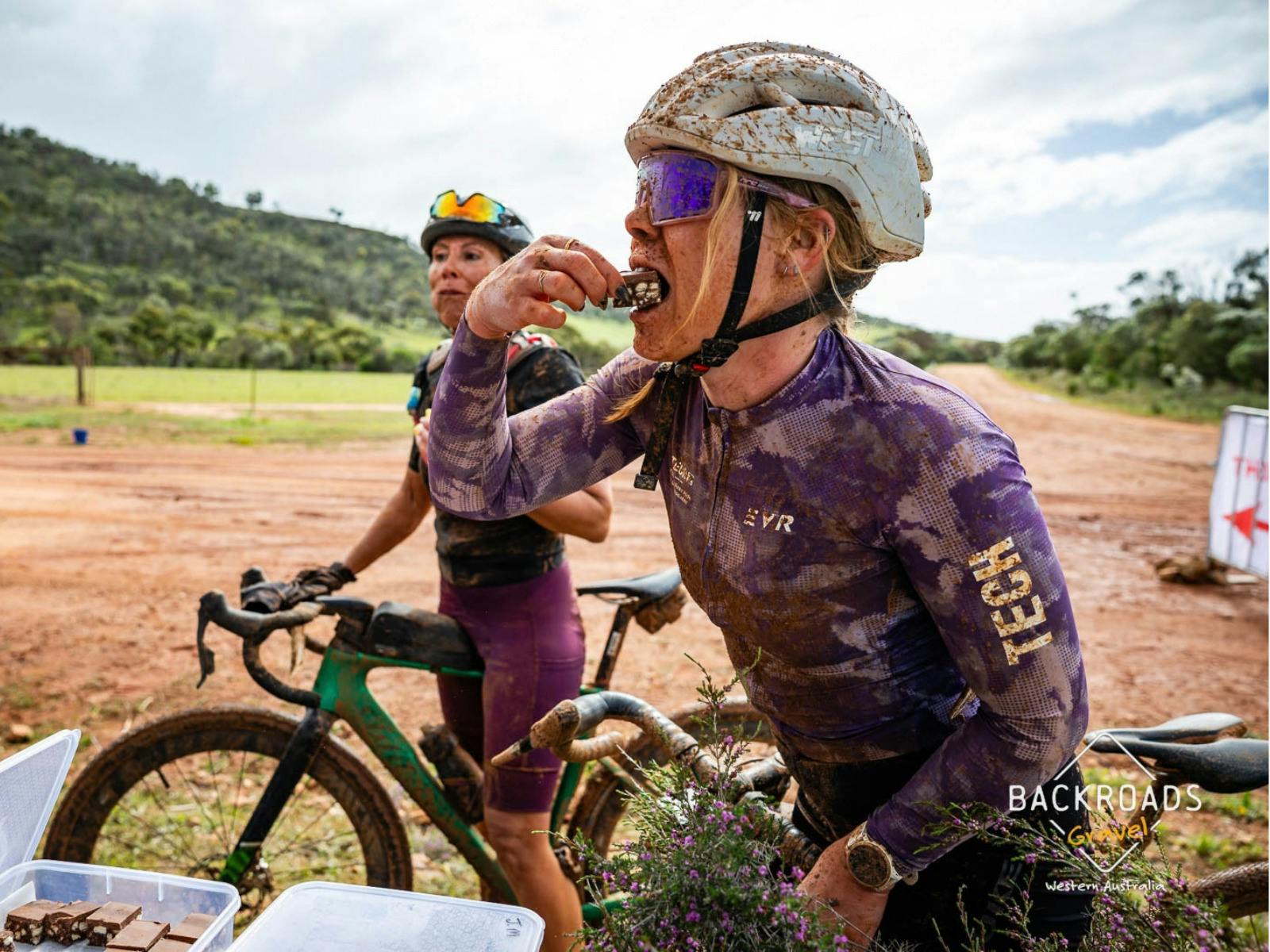 Mud-covered cyclist eating at an aid station during Backroads Gravel.