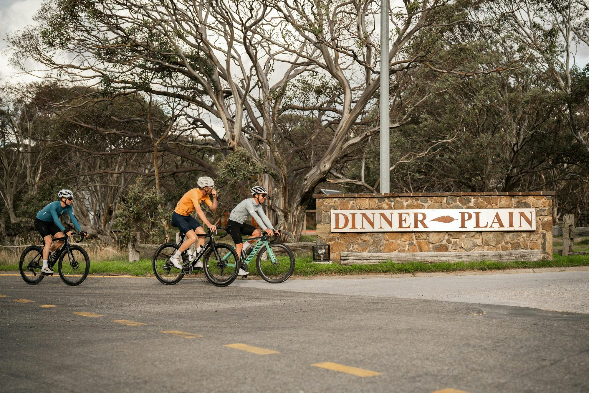 Three cyclists riding past the Dinner Plain sign on the 7 Peaks Challenge route.