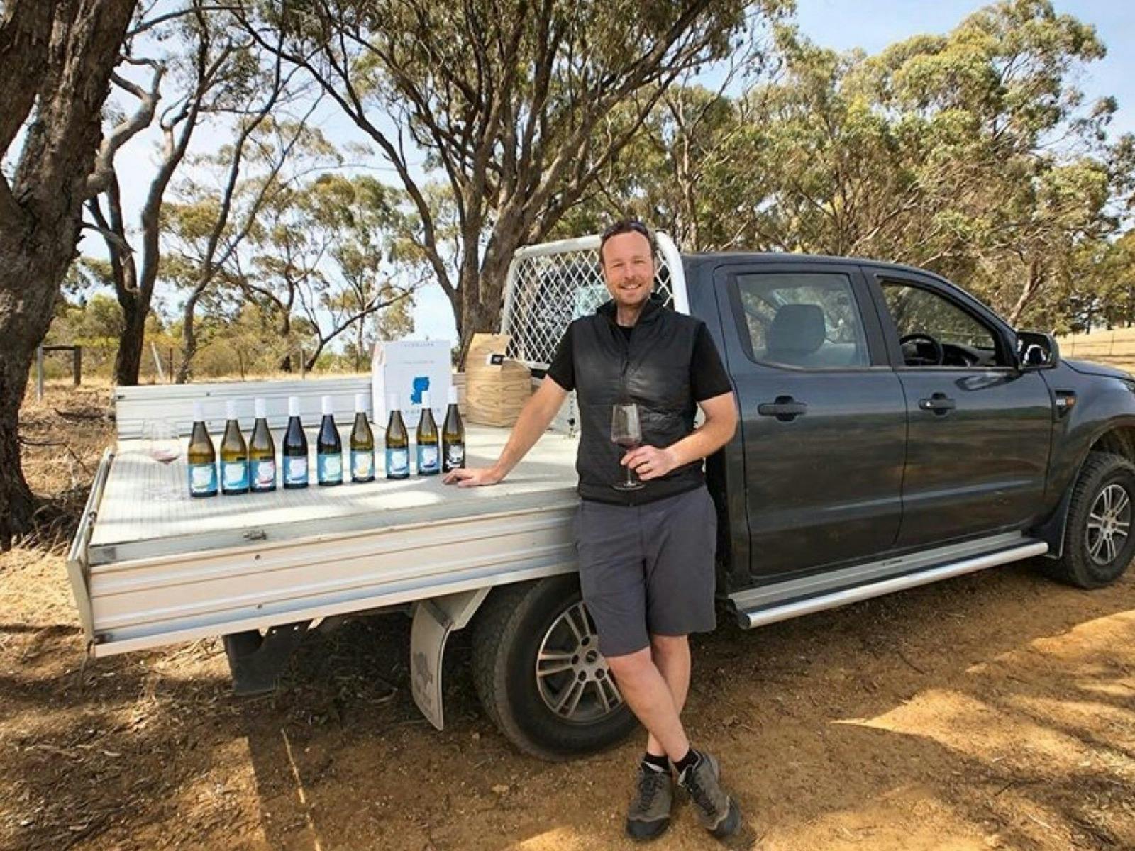 A man stands leaning on a work ute adorned with a selection of premium wine bottles