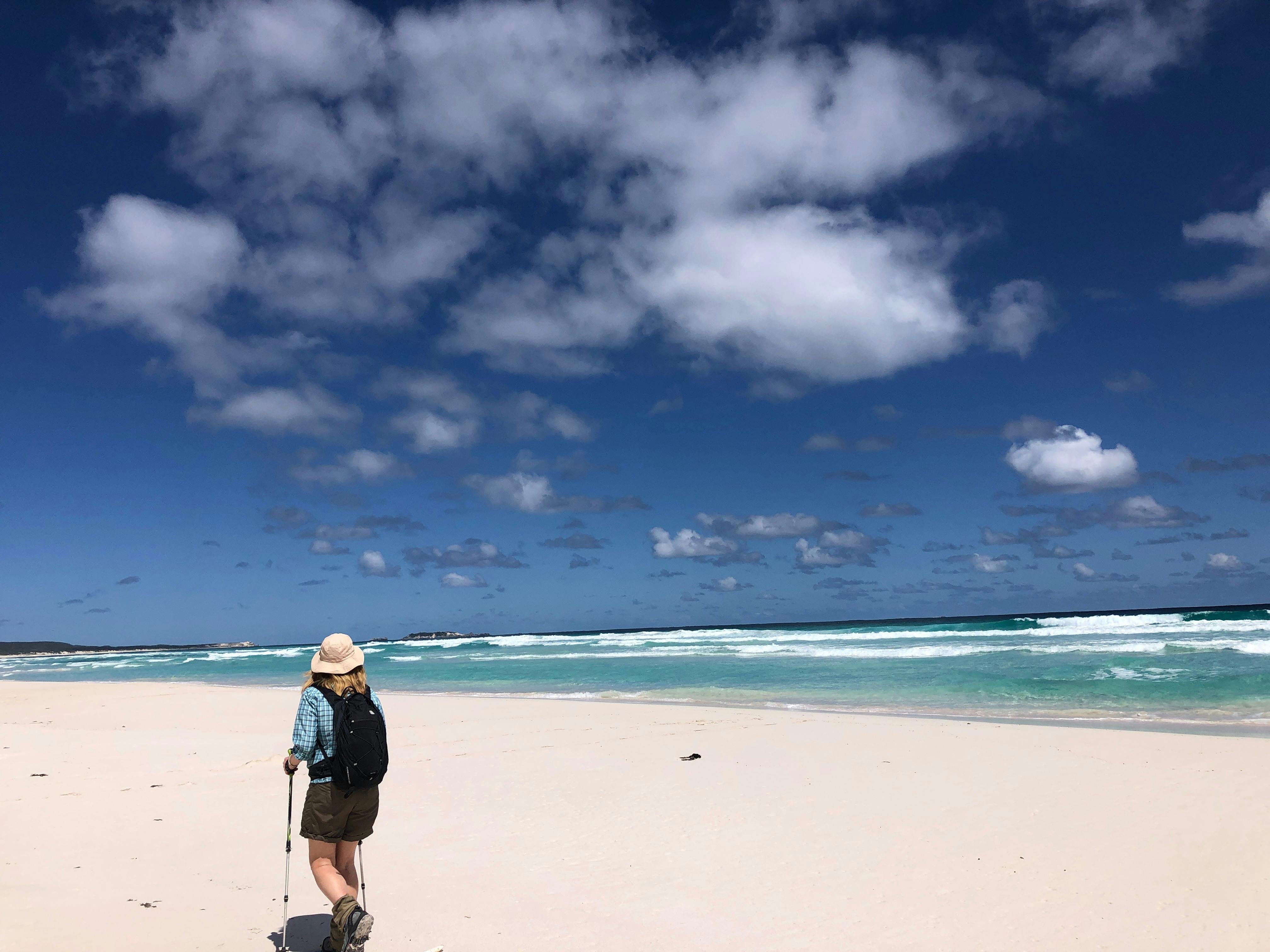 a woman with walking poles walking along the beach in front of her bright blue sky and ocean.
