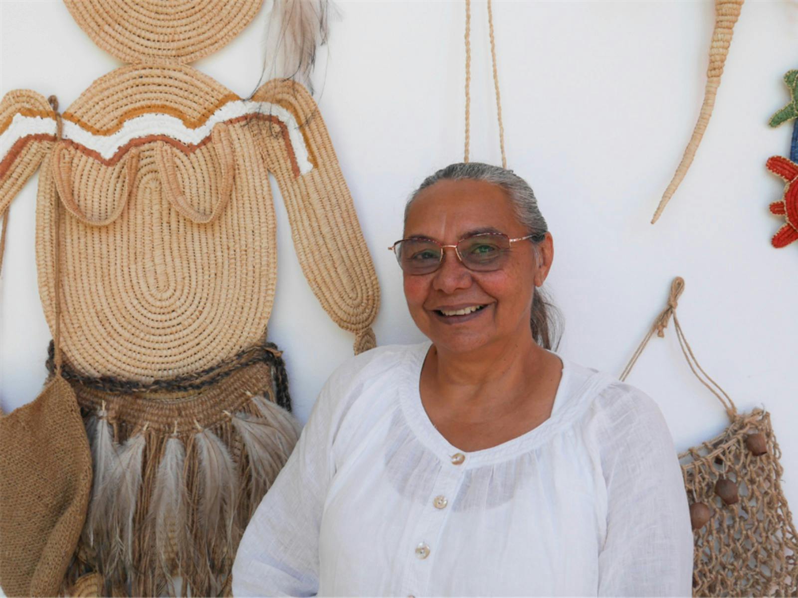 Photograph of Aunty Annette standing in front of her weaving artworks that are hung on a white wall.