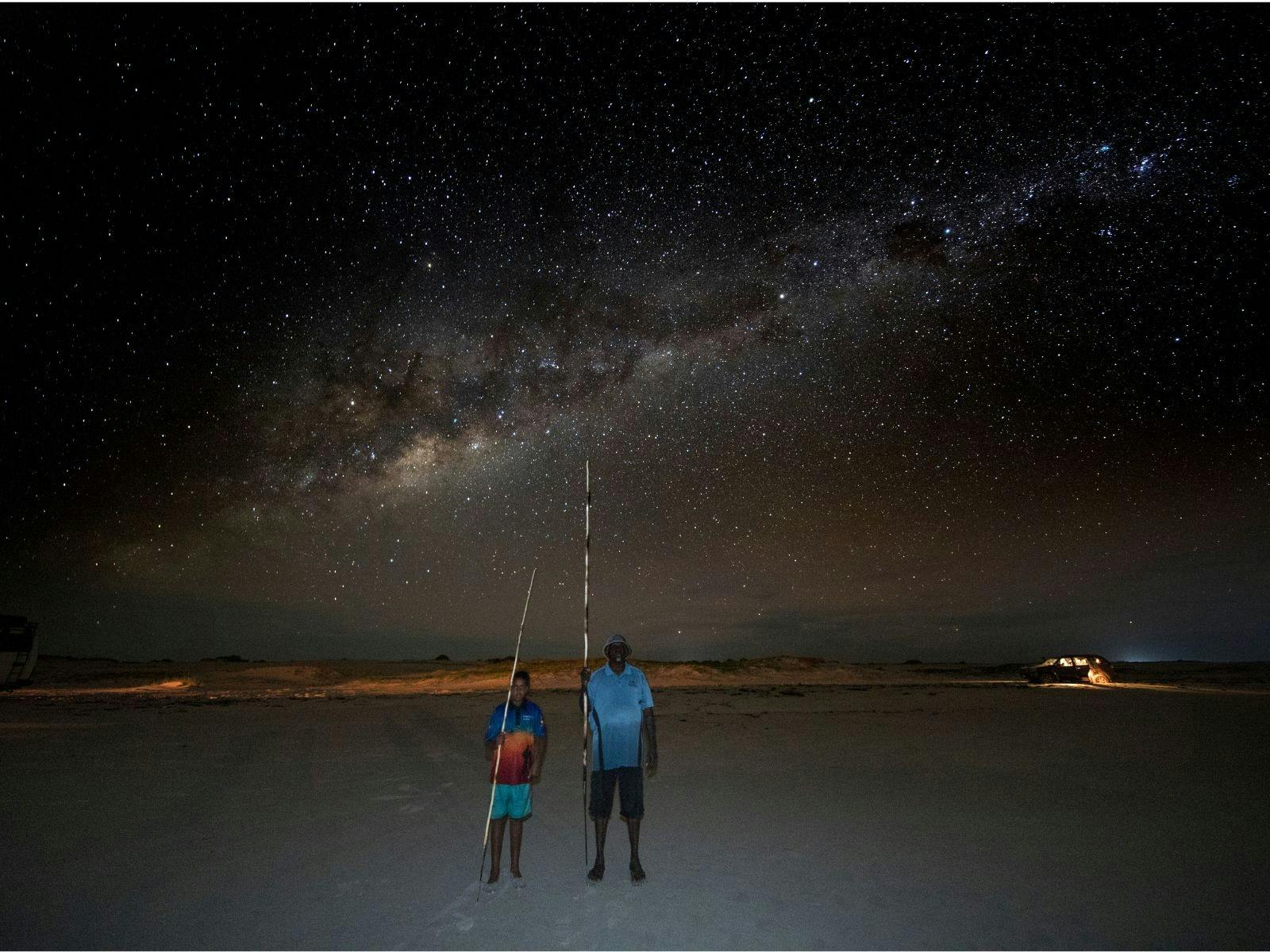 Bundy and boy standing on beach with their spears below the milky way.
